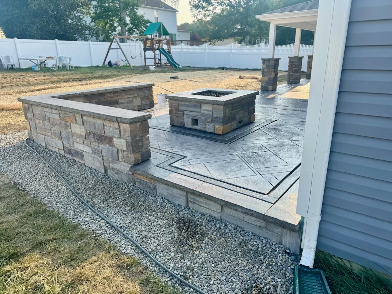 Stone patio with built-in wall and fire pit, with a lawn and play set in the background.