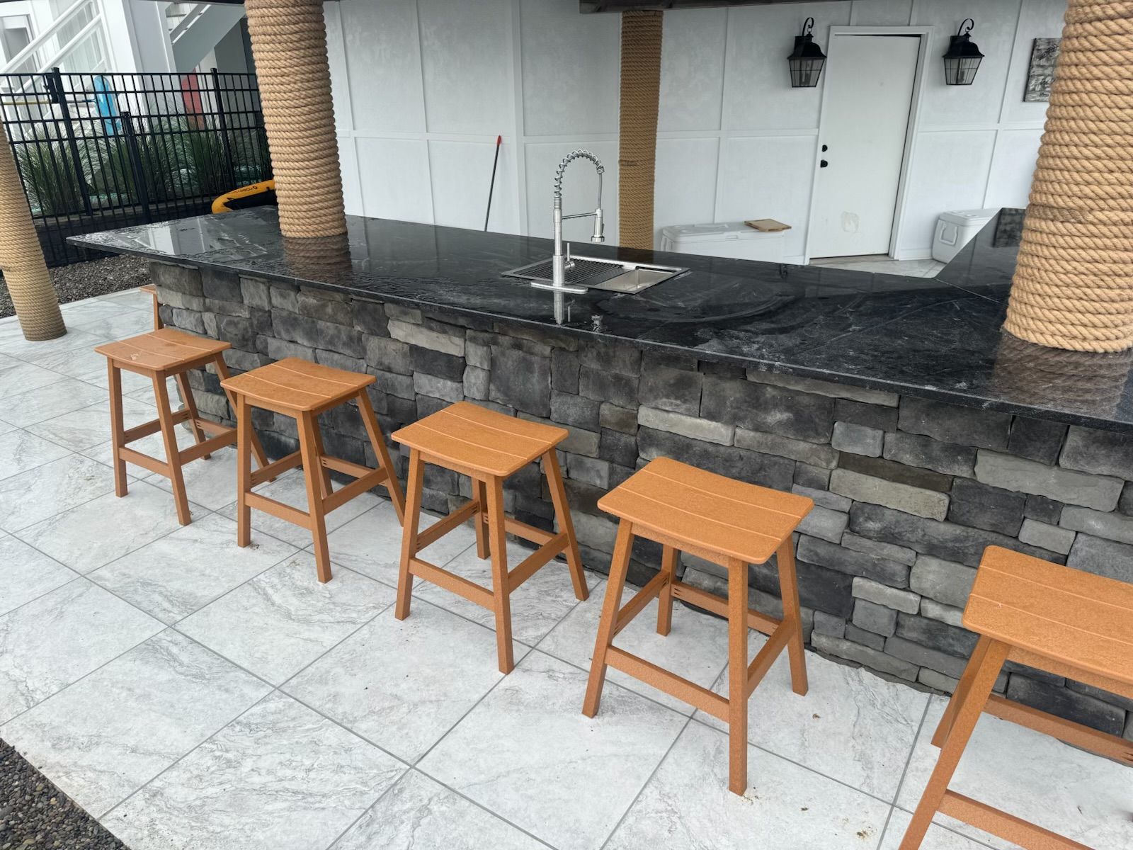 Outdoor bar with stone facade, black countertop, wooden stools, and rope-wrapped pillars.