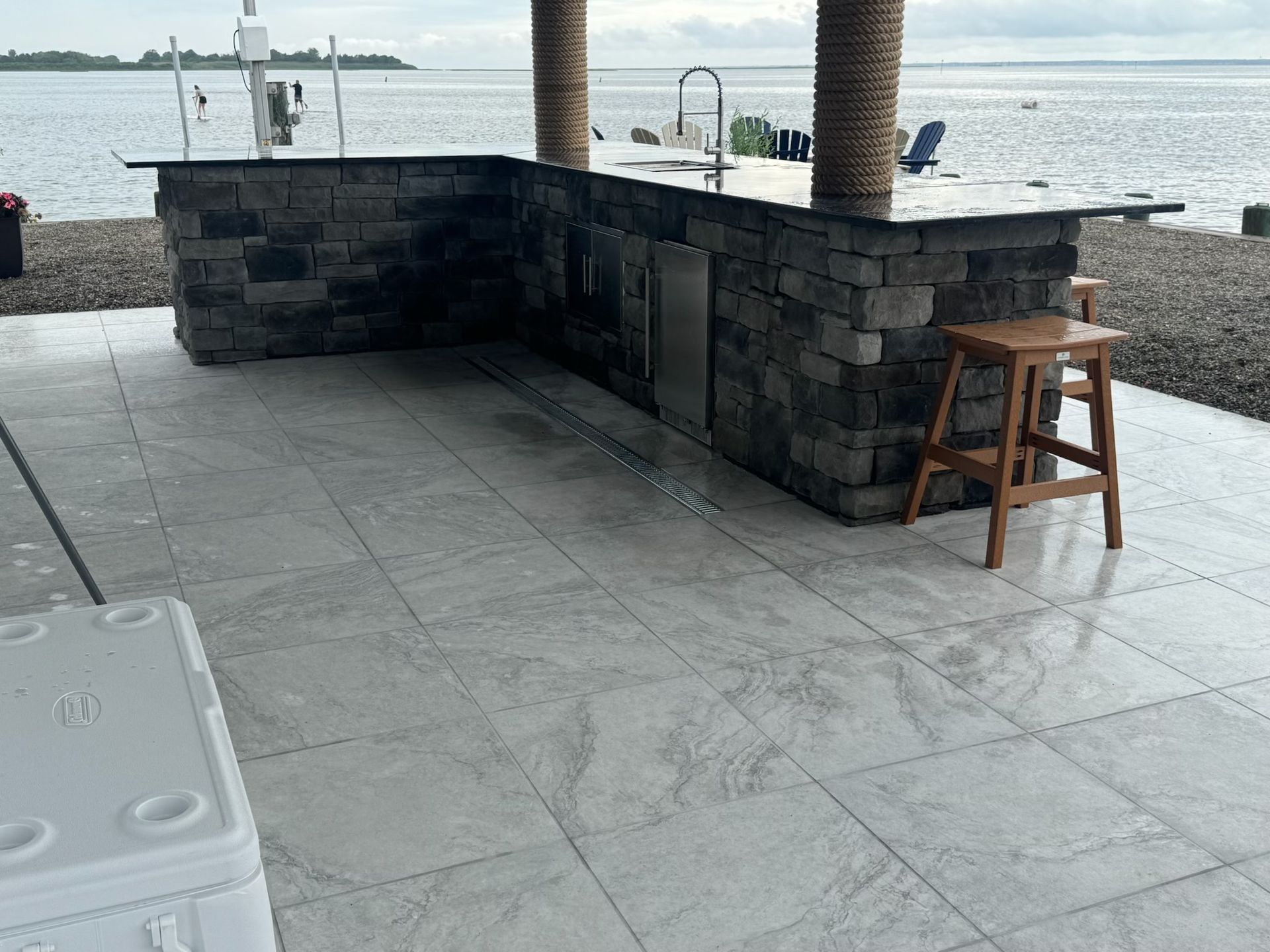 Outdoor bar with stone facade, gray tile floor, water view, and wooden bar stool.