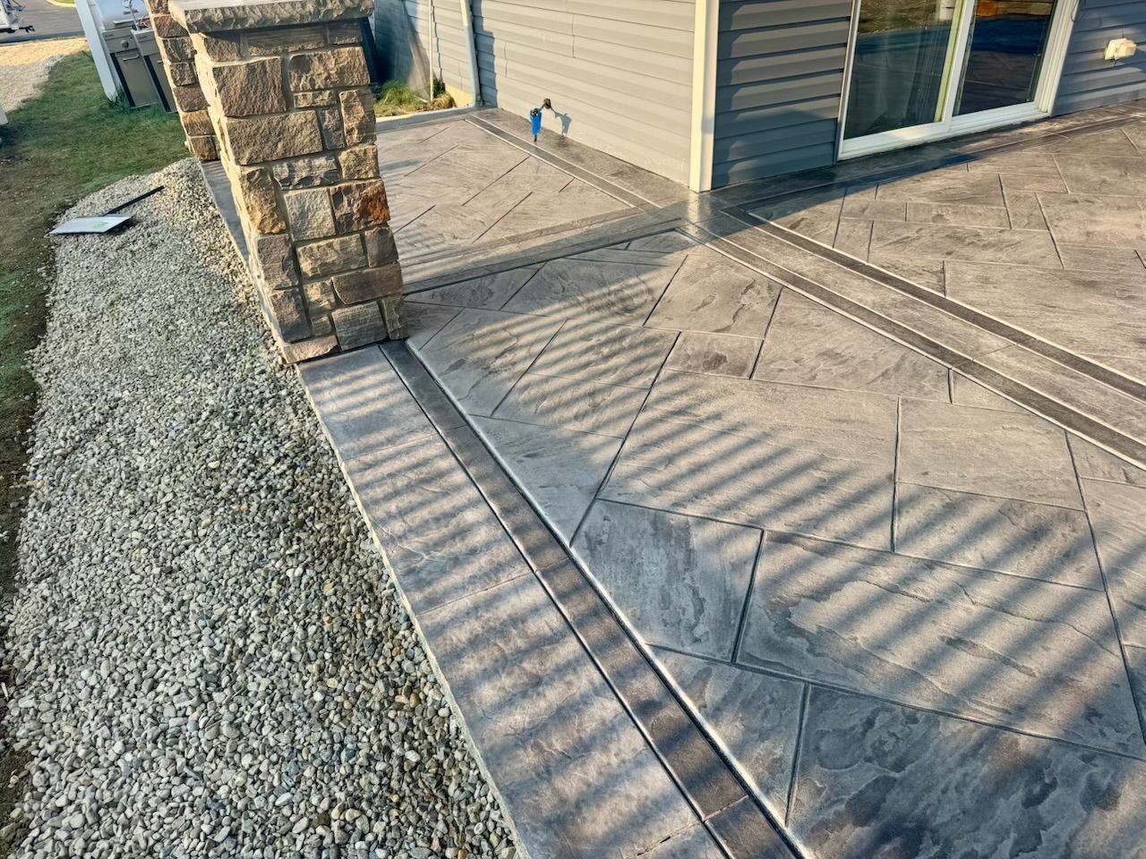 A gray stamped concrete patio with a decorative border next to a gravel and grass lawn.