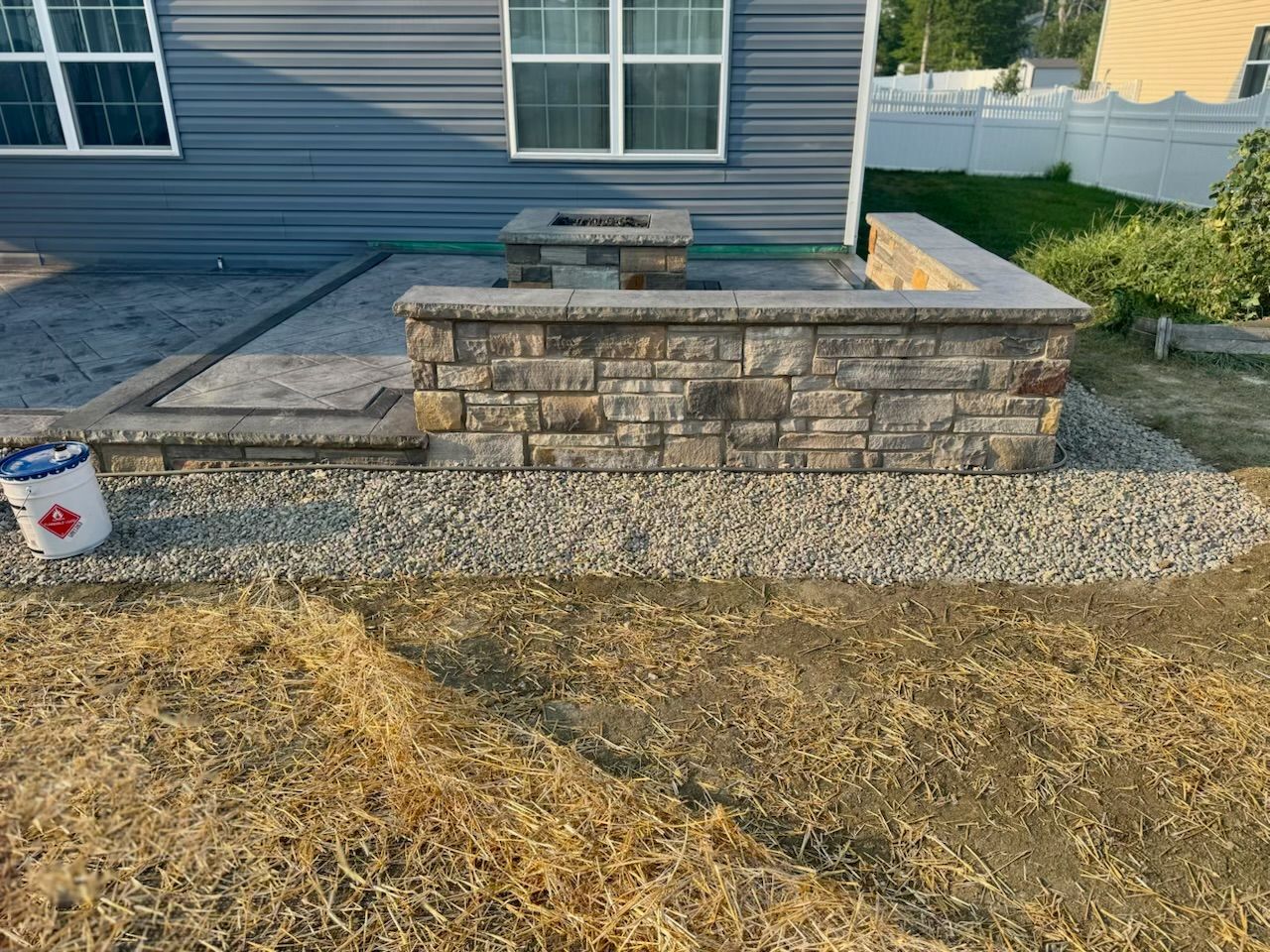 Stone patio with a fire pit and retaining wall. Gray siding and gravel surround.