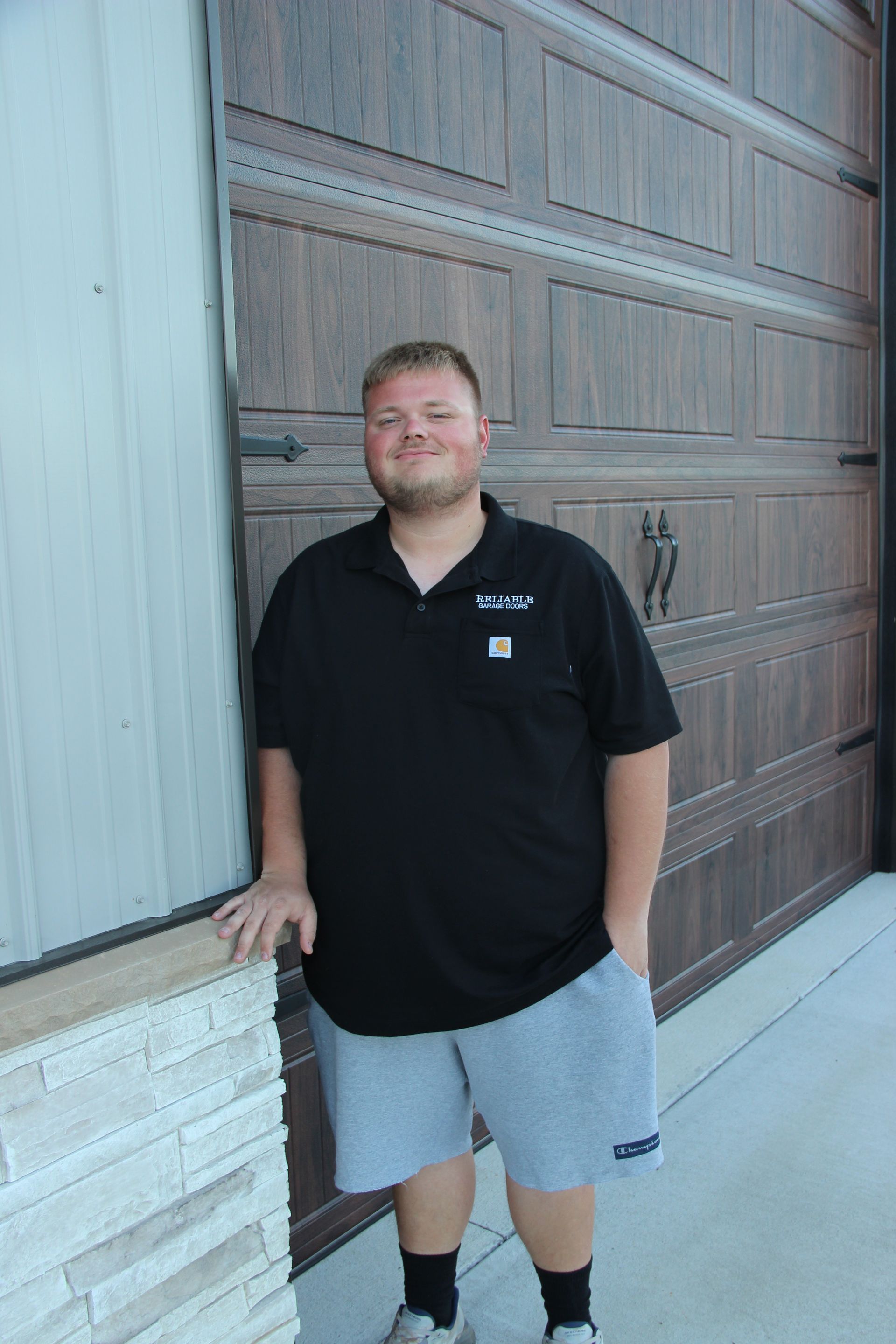 Man standing by a large wooden door, wearing a black polo and gray shorts, smiling at the camera.