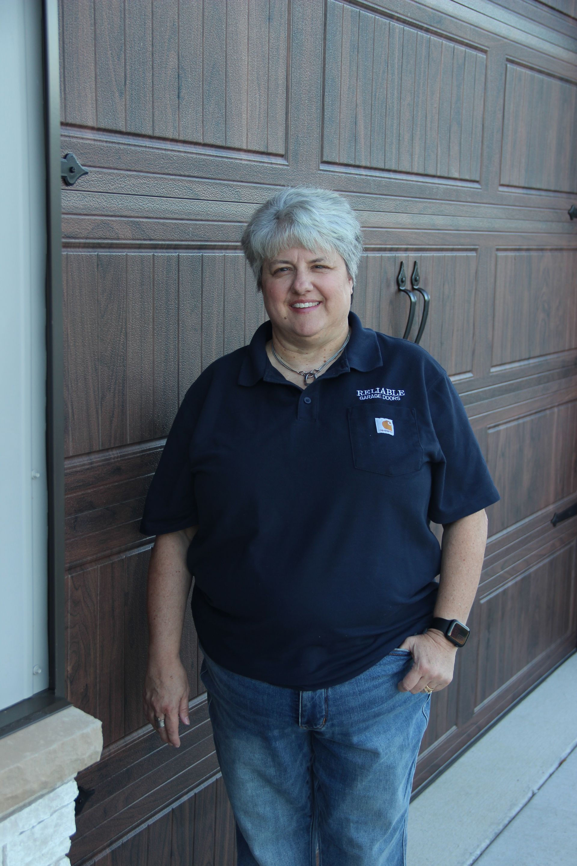 Smiling person in a navy polo and jeans standing beside a wooden garage door