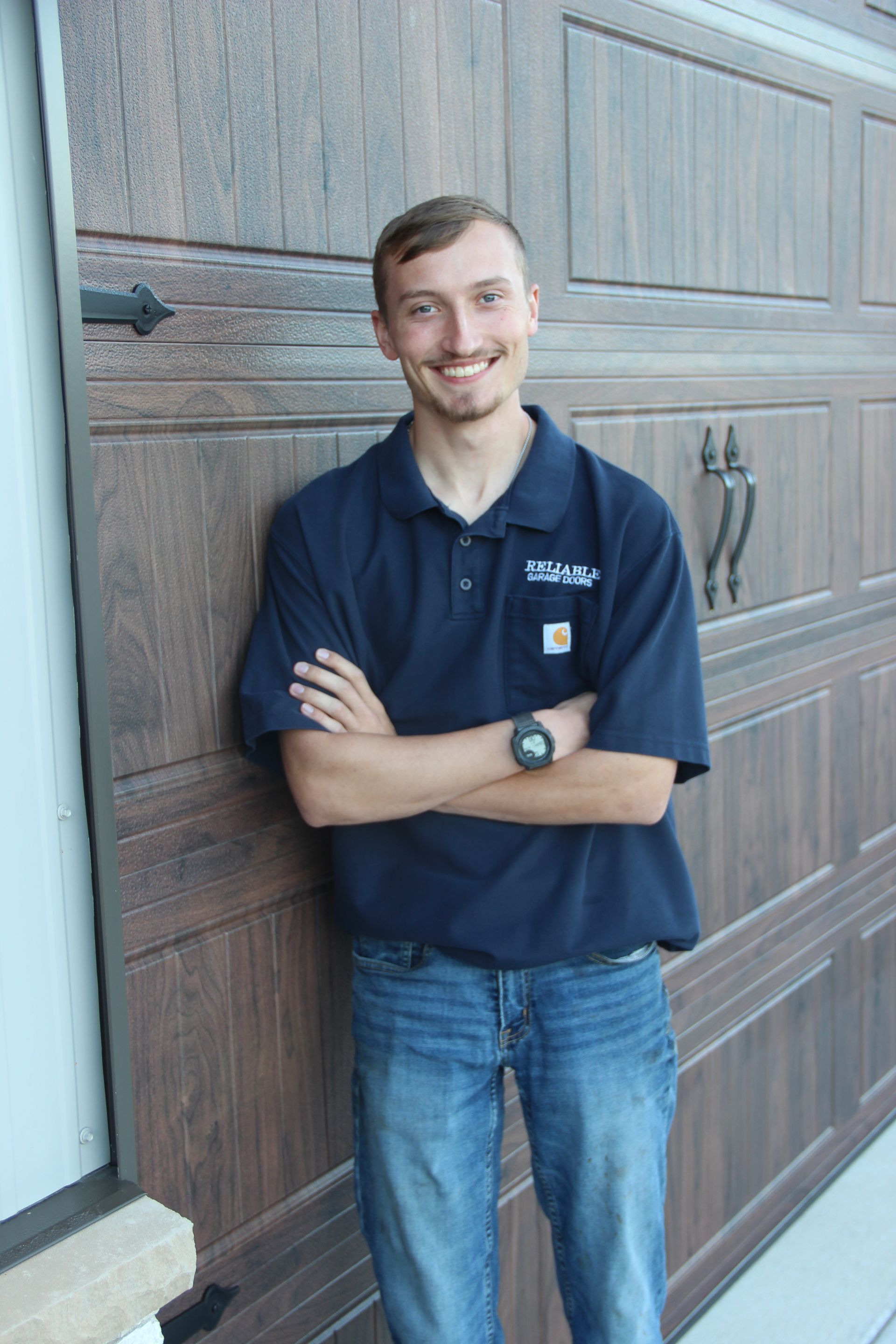 Smiling person in a navy polo shirt and jeans leaning against a wooden garage door, arms crossed.