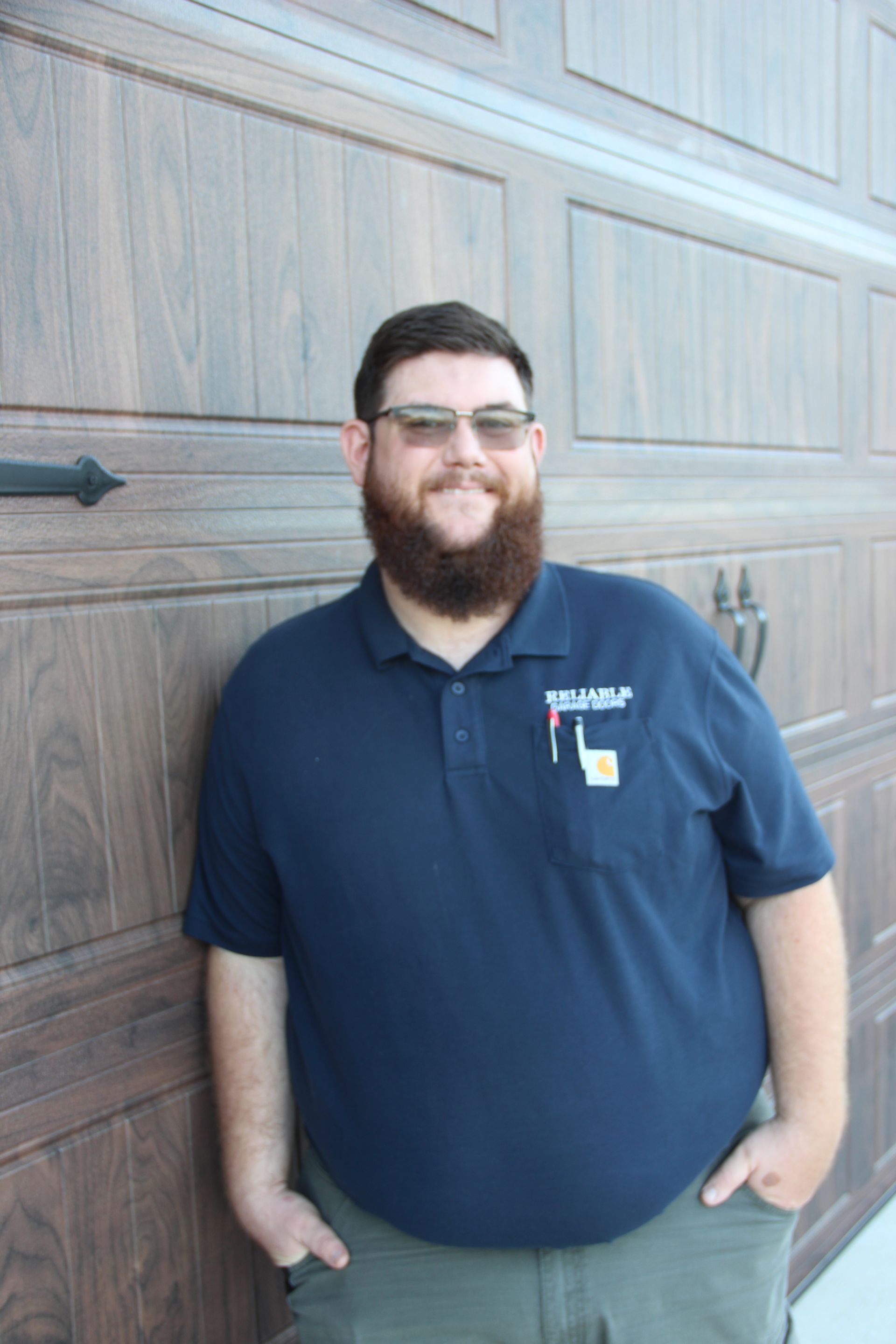 Man standing in front of a garage door, wearing glasses, a beard, and a navy polo shirt, smiling with hands in pockets