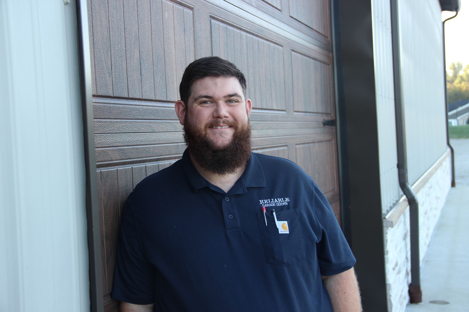 Smiling man in a navy polo shirt standing beside a wooden wall outdoors