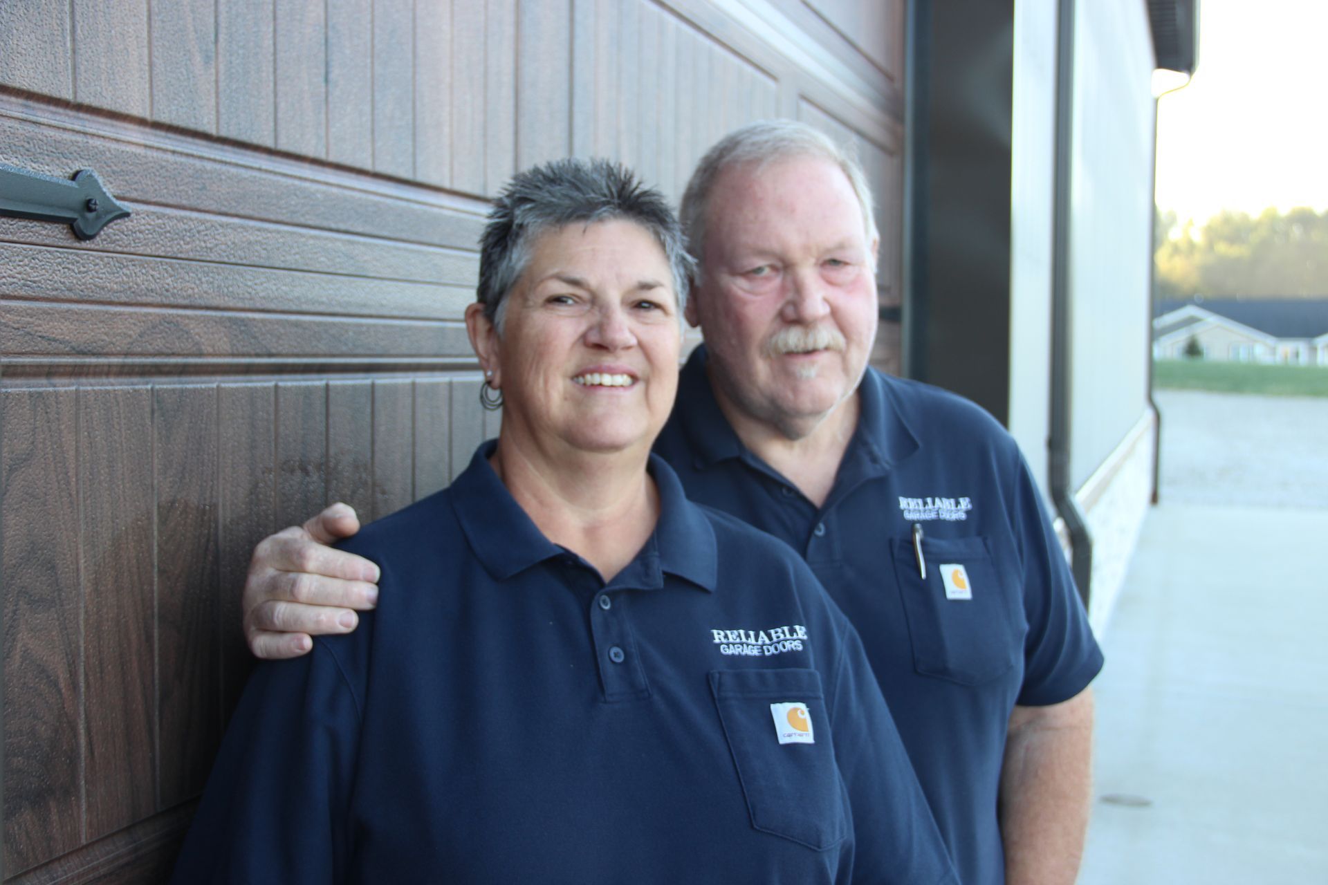 Two people in navy polos standing by a wooden wall, smiling at the camera.