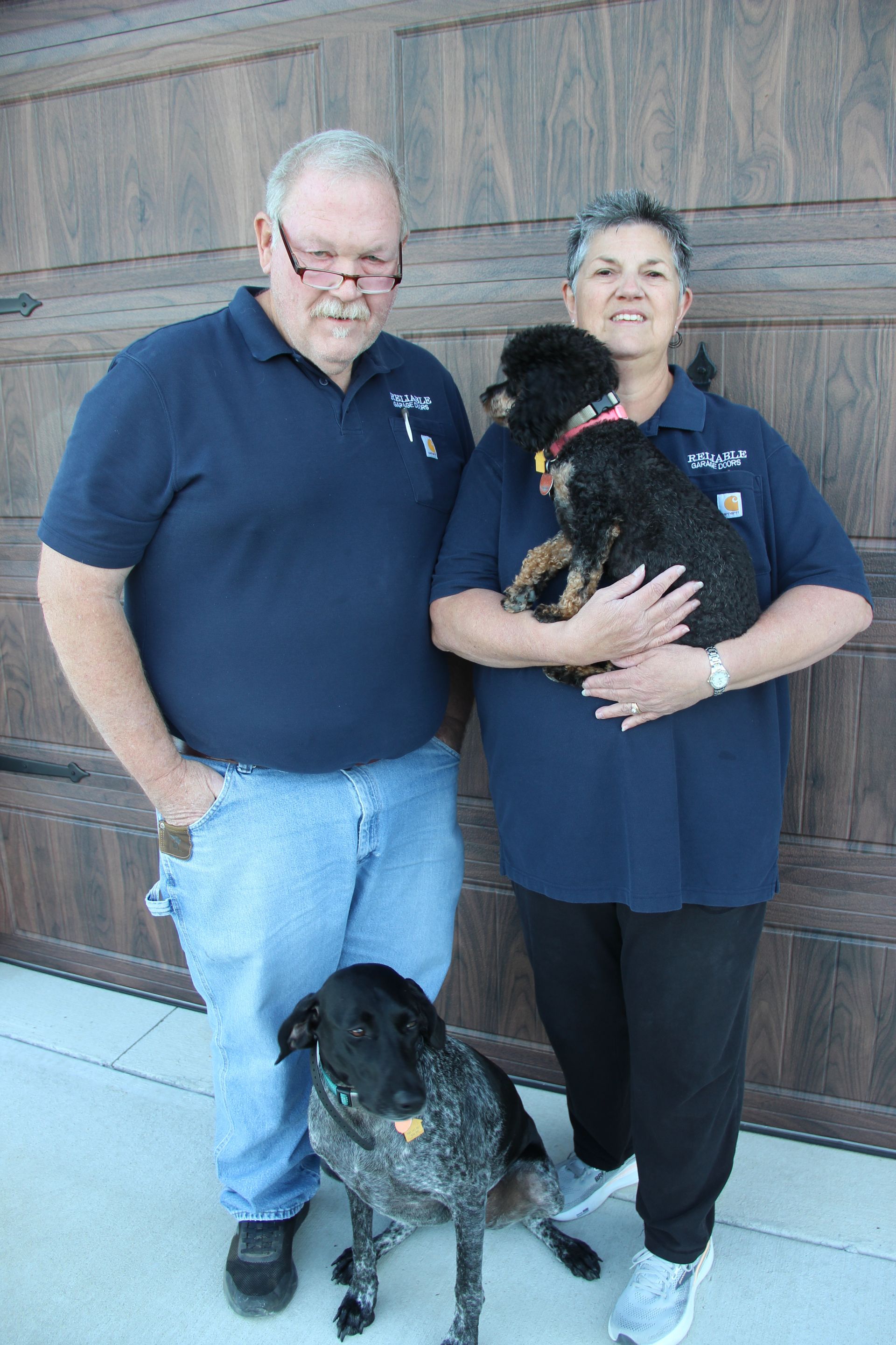Two people in navy shirts standing by a wooden door, holding a small dog, with a larger dog at their feet.