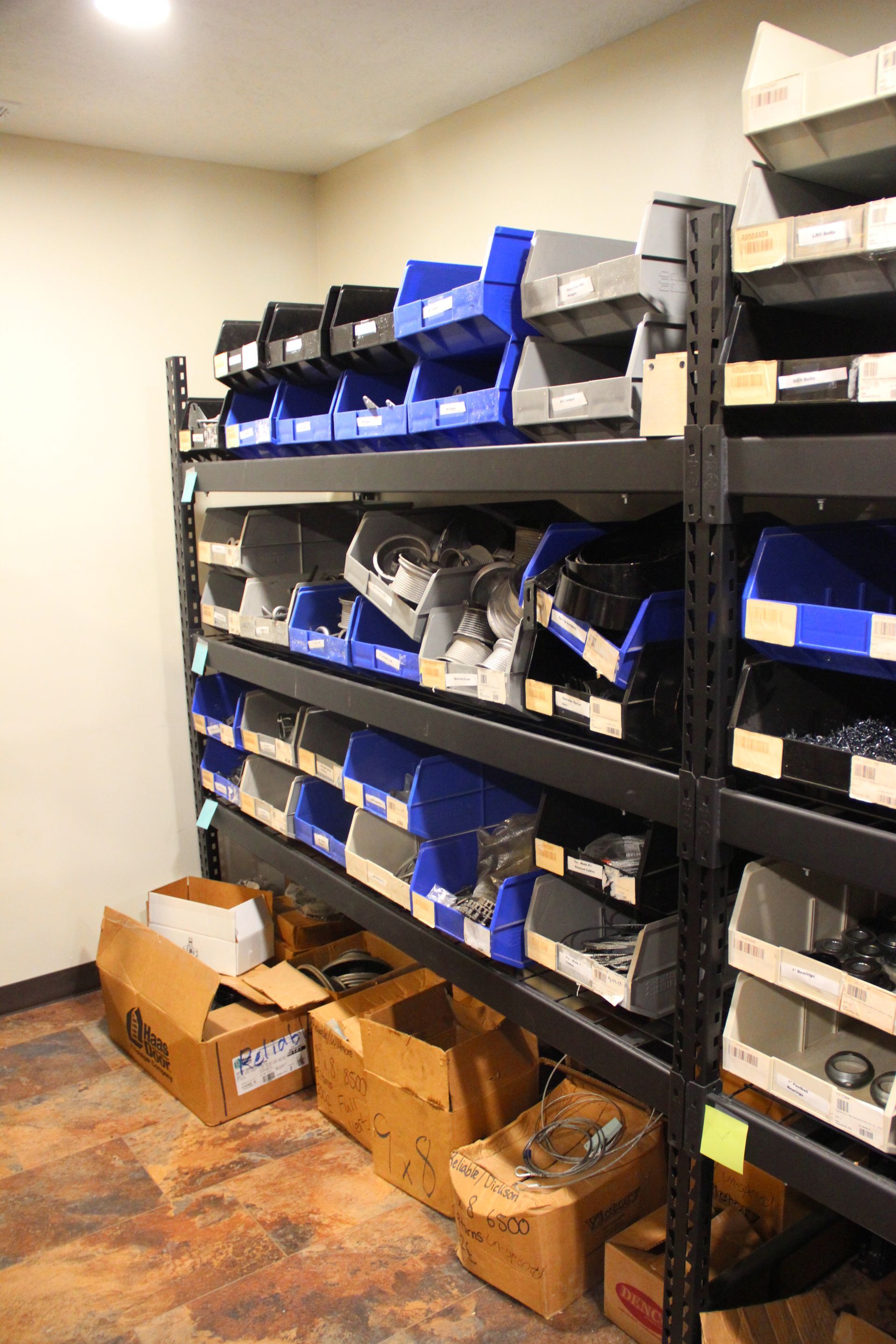 Shelves of labeled storage bins in a small supply room, with cardboard boxes on the floor
