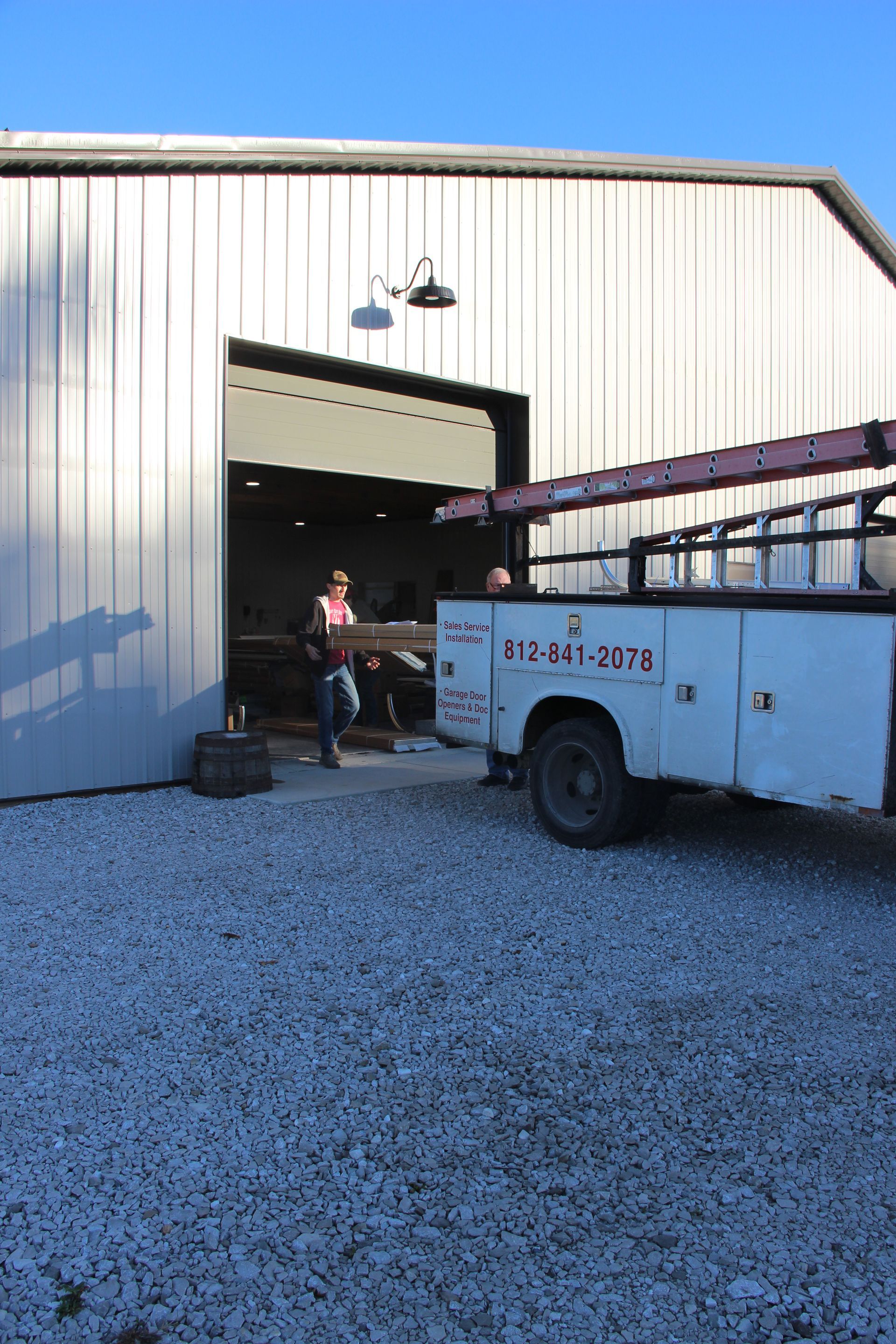Person standing by a white utility truck outside a large metal warehouse entrance.