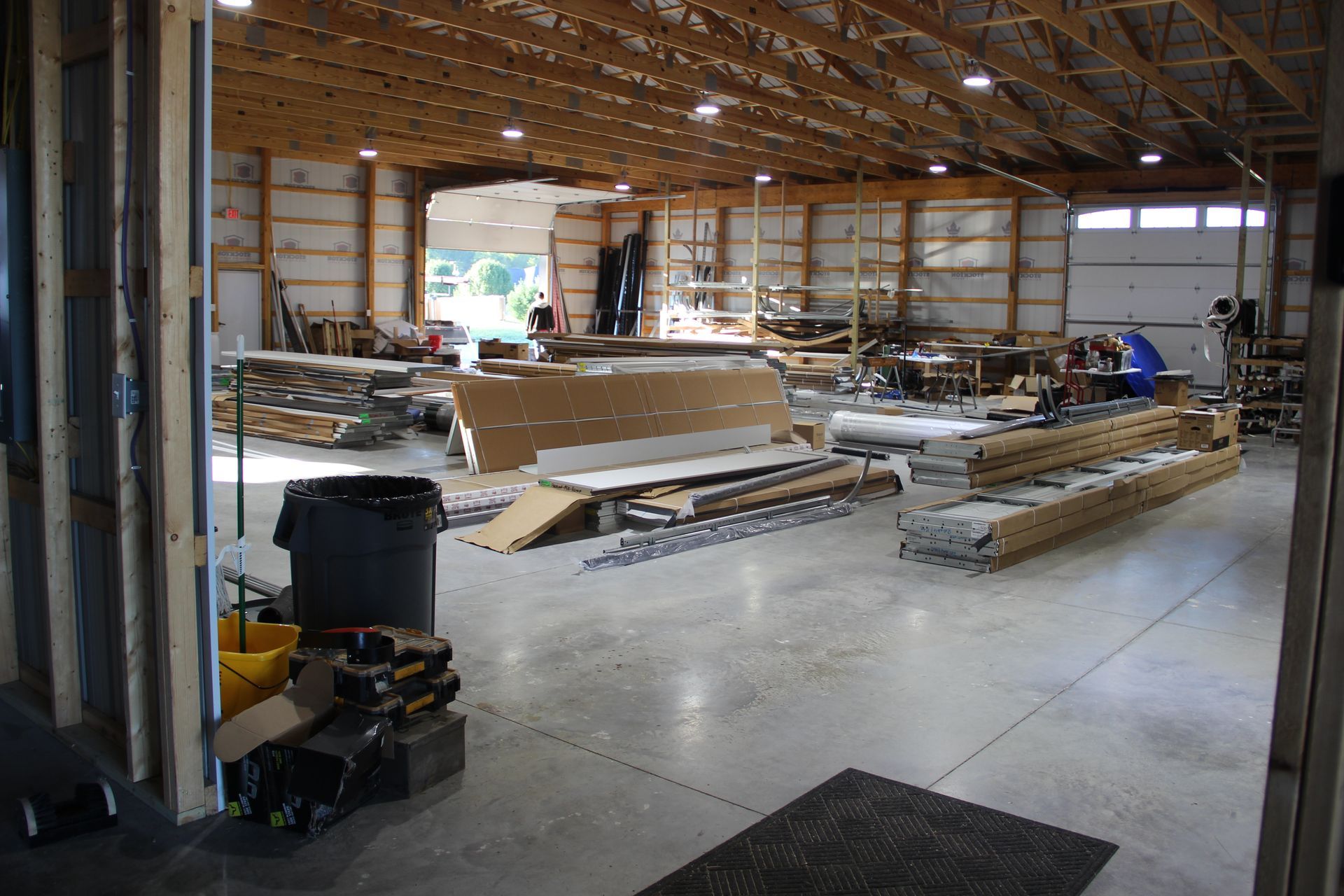 Interior of a warehouse with lumber, stacked materials, and a concrete floor under a wooden roof.