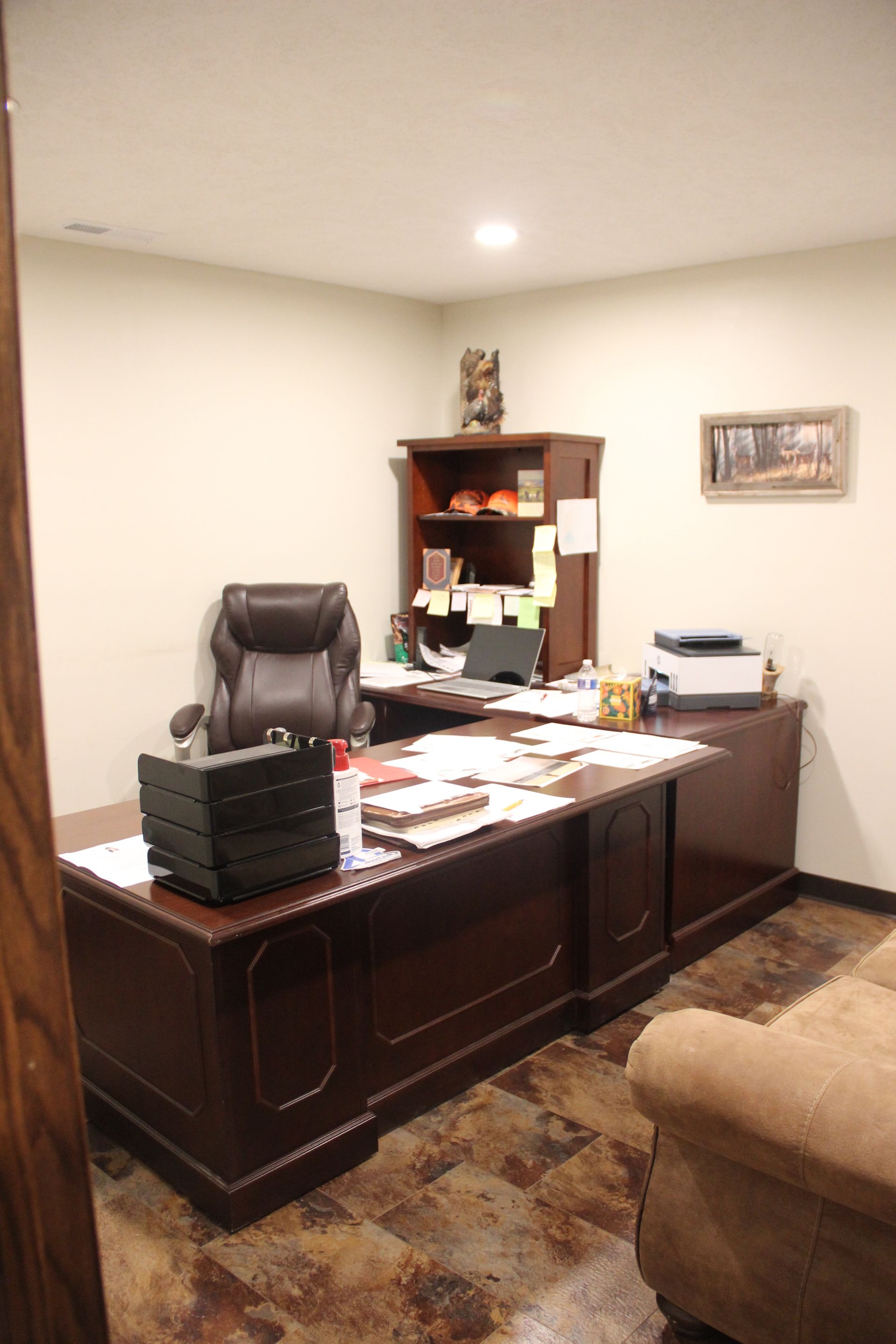 Office with dark wood desk, leather chair, bookshelves, lamp, and beige sofa in the corner.