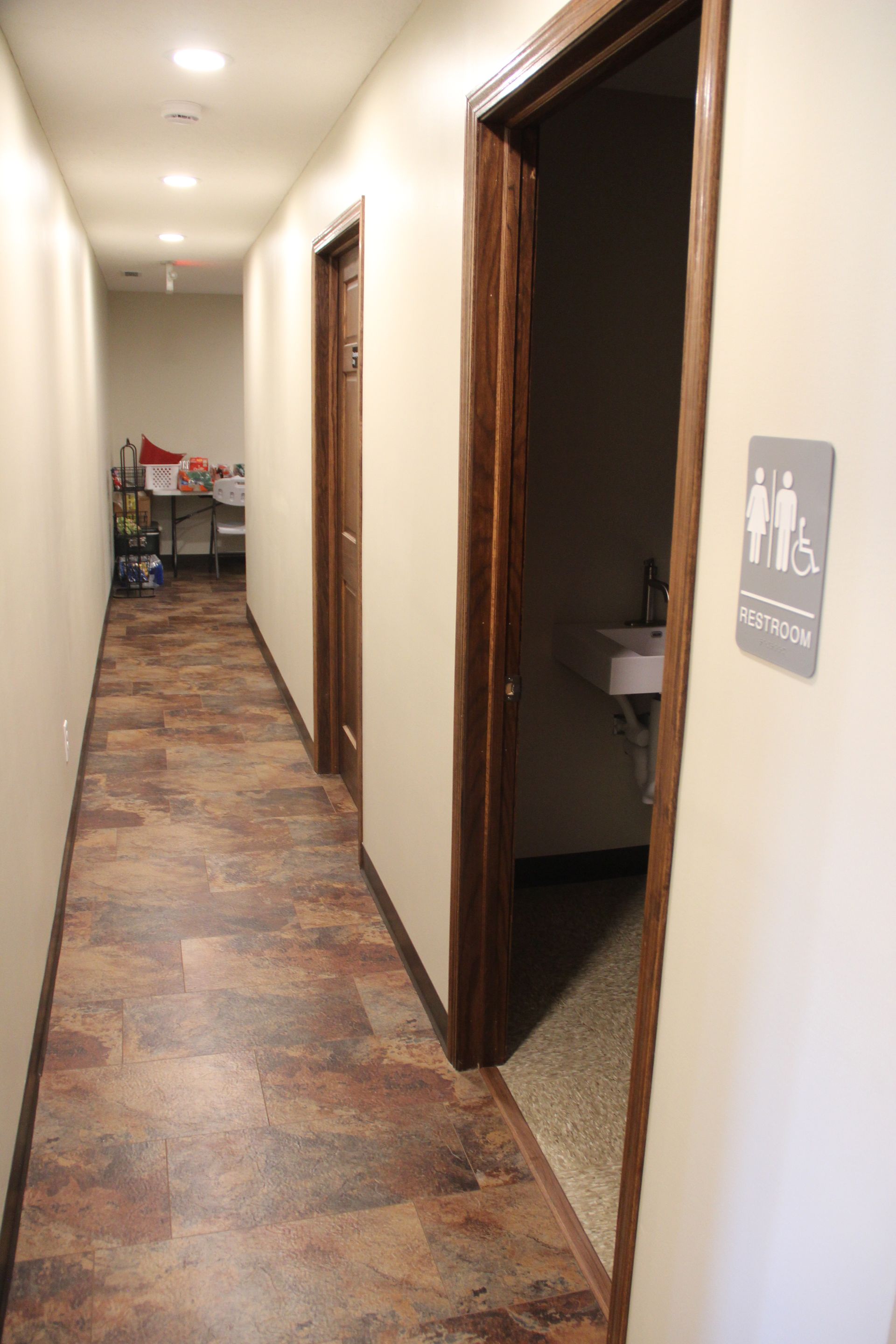Narrow hallway with doors and restroom sign, carpeted floor, and seating visible at the end.
