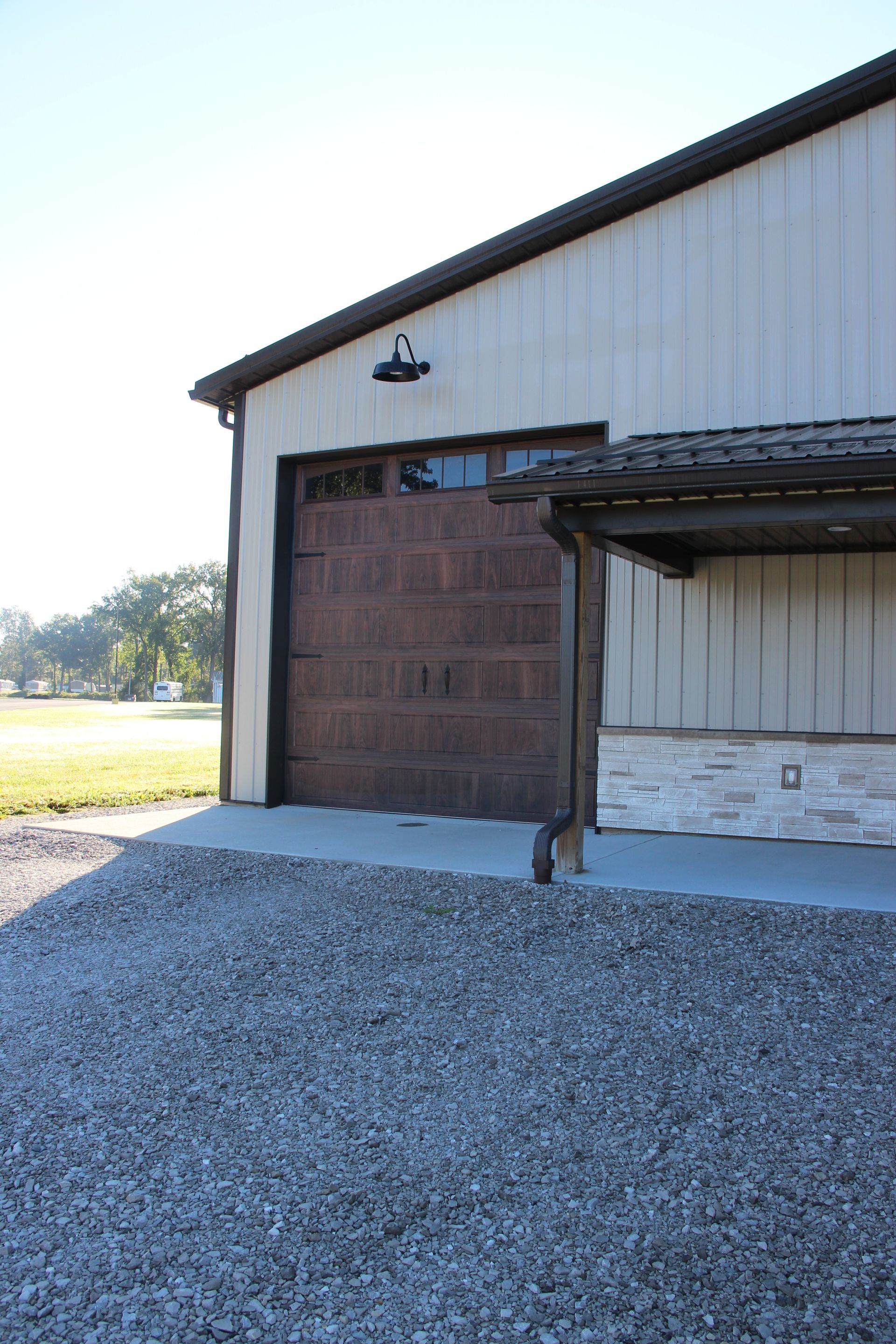 Metal building with a large wooden garage door and gravel driveway beside a grassy field.