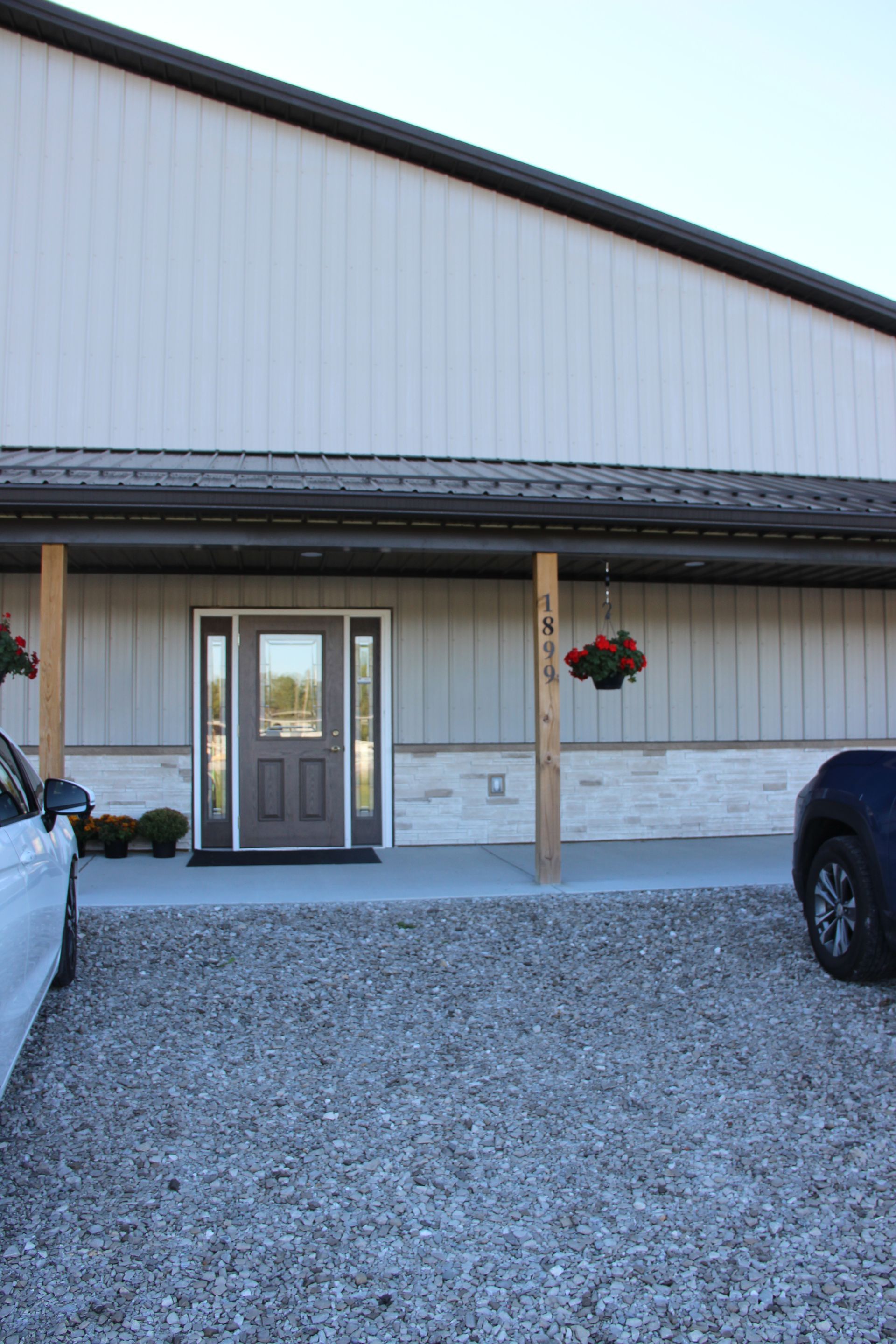 Gray building entrance with a covered porch, hanging flower basket, and parked cars on a gravel lot