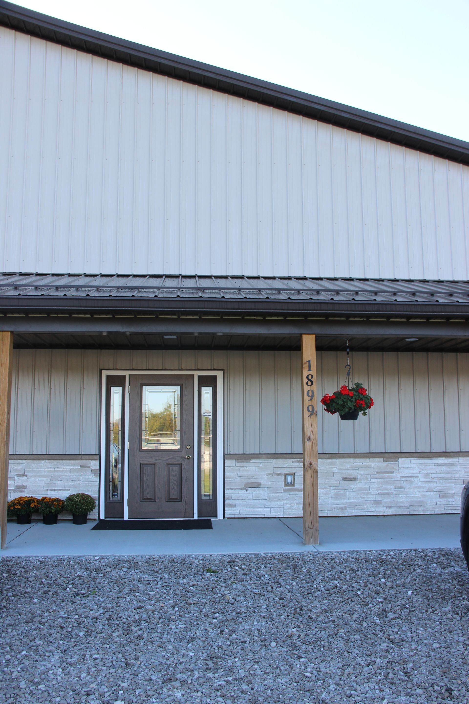 Front entrance of a metal-sided building with double glass doors, wood post, hanging flowers, and gravel foreground