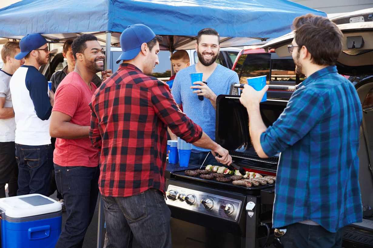 A group of friends gathers for a backyard barbecue, grilling food and holding drinks under a blue canopy.