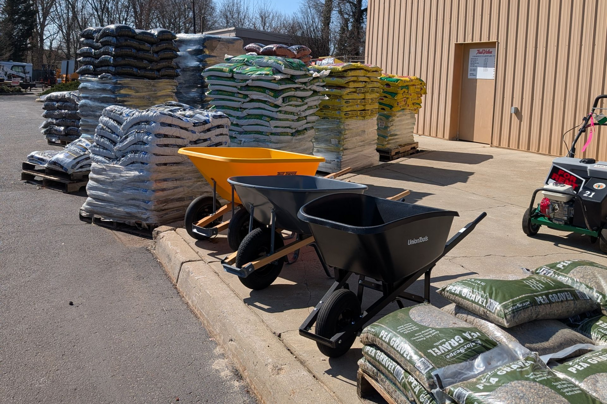 Stacked bags of landscaping material and three wheelbarrows—yellow, gray, and black—sit on a concrete lot.