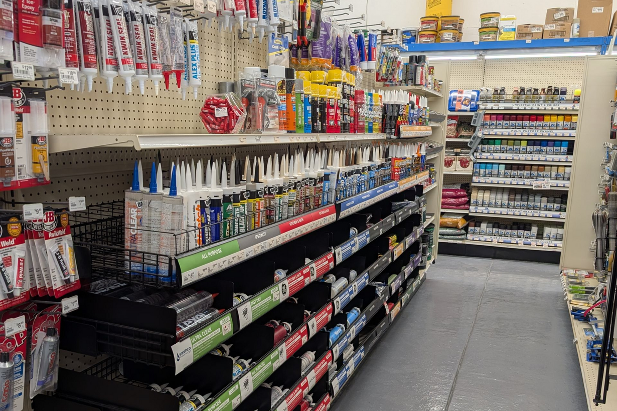 A view down a hardware store aisle featuring shelves stocked with various tubes of sealant, caulk, and spray paint.