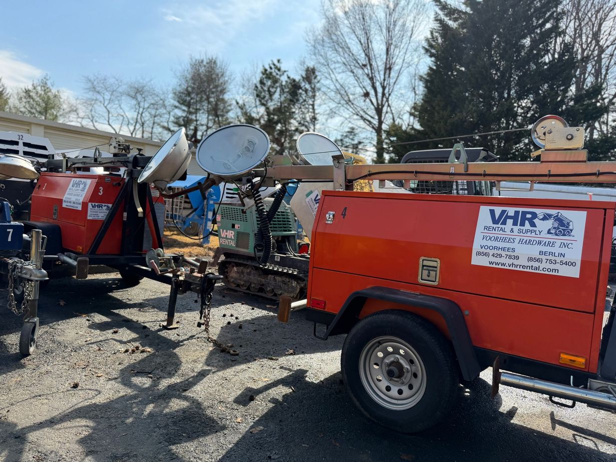 Bright orange VHR utility trailers with mounted floodlights parked on a gravel lot under a clear blue sky.