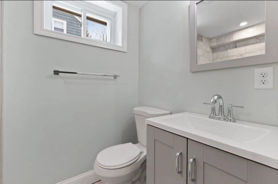 A bathroom with a toilet, sink, mirror and window.