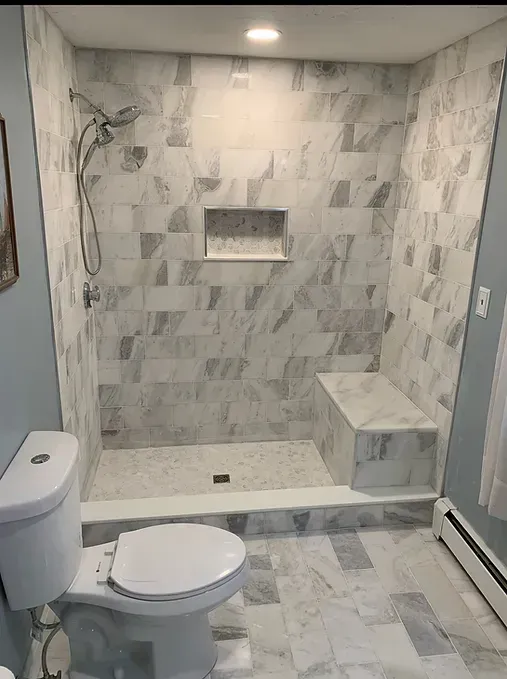 A bathroom with a toilet , shower , and bench.