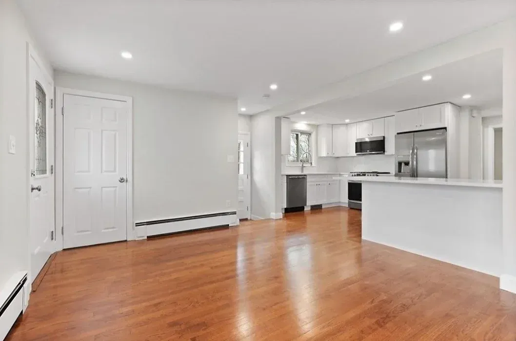 An empty living room with hardwood floors and a kitchen in the background.