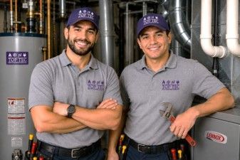Two men in gray uniforms and purple caps smiling in an industrial room with pipes and equipment