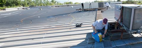 Workers on a rooftop, one kneeling by tools near a white truck under a bright sky.