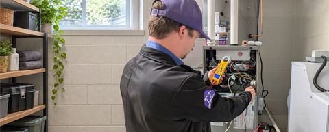 HVAC technician inspecting furnace with a multimeter, indoors. Wearing a purple hat, black jacket.
