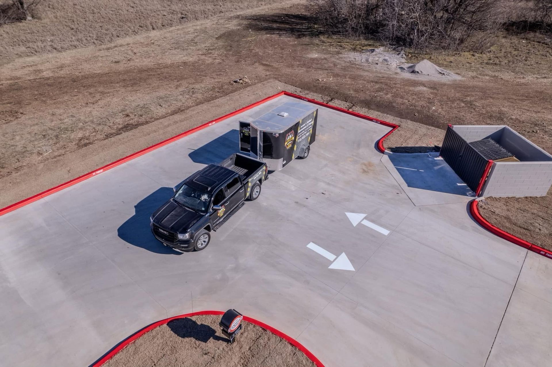Black pickup truck towing a silver trailer on a concrete lot with red curbs and white arrows.