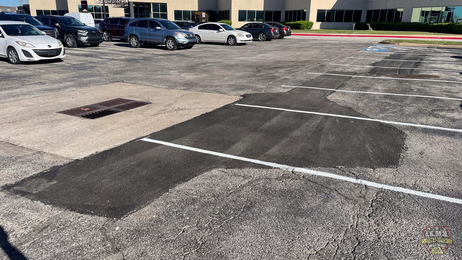 Mostly empty parking lot with several parked cars and worn asphalt striping in front of a building.