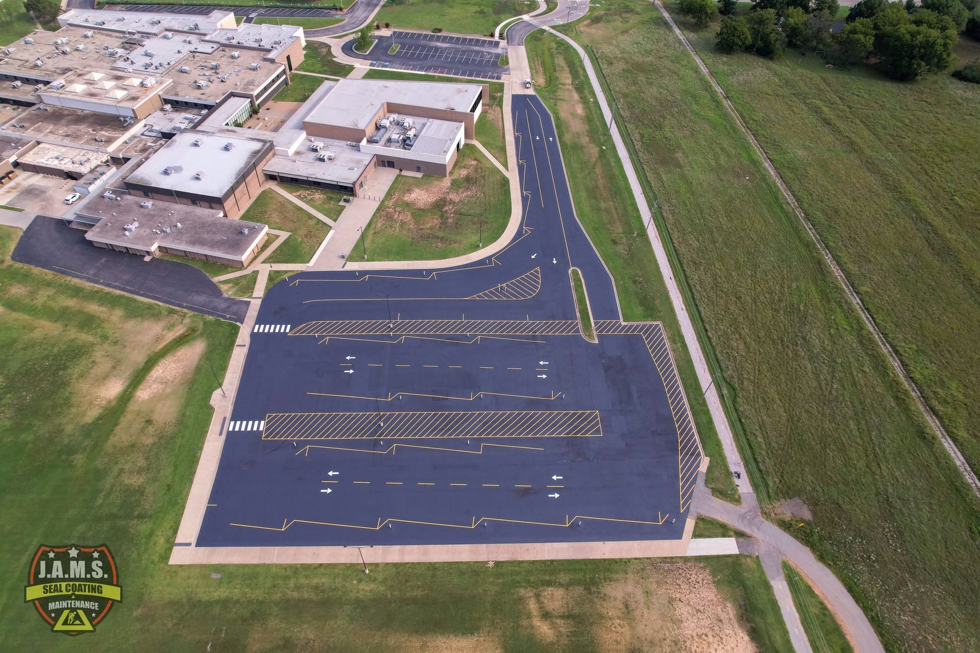 Aerial view of a school parking lot beside buildings and fields, with fresh asphalt and painted lanes.