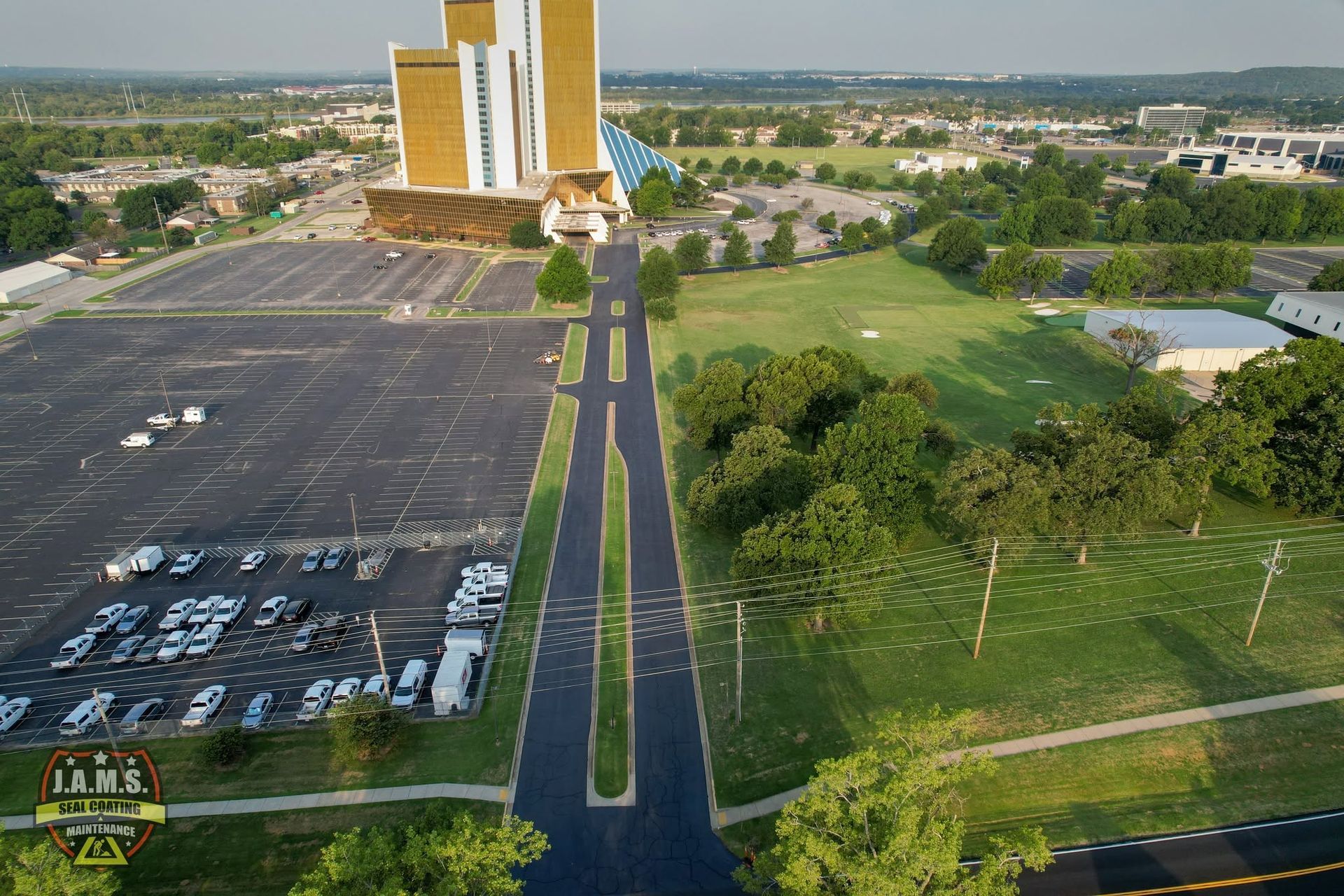 Aerial view of a campus entrance road between parking lots and green lawns, with tall gold towers ahead.