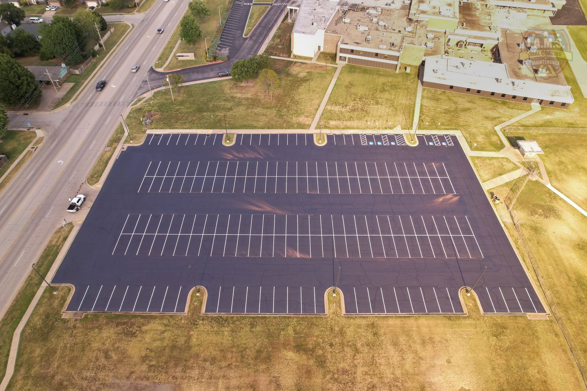 Aerial view of an empty parking lot beside a road and buildings, with white lined spaces and a dark surface