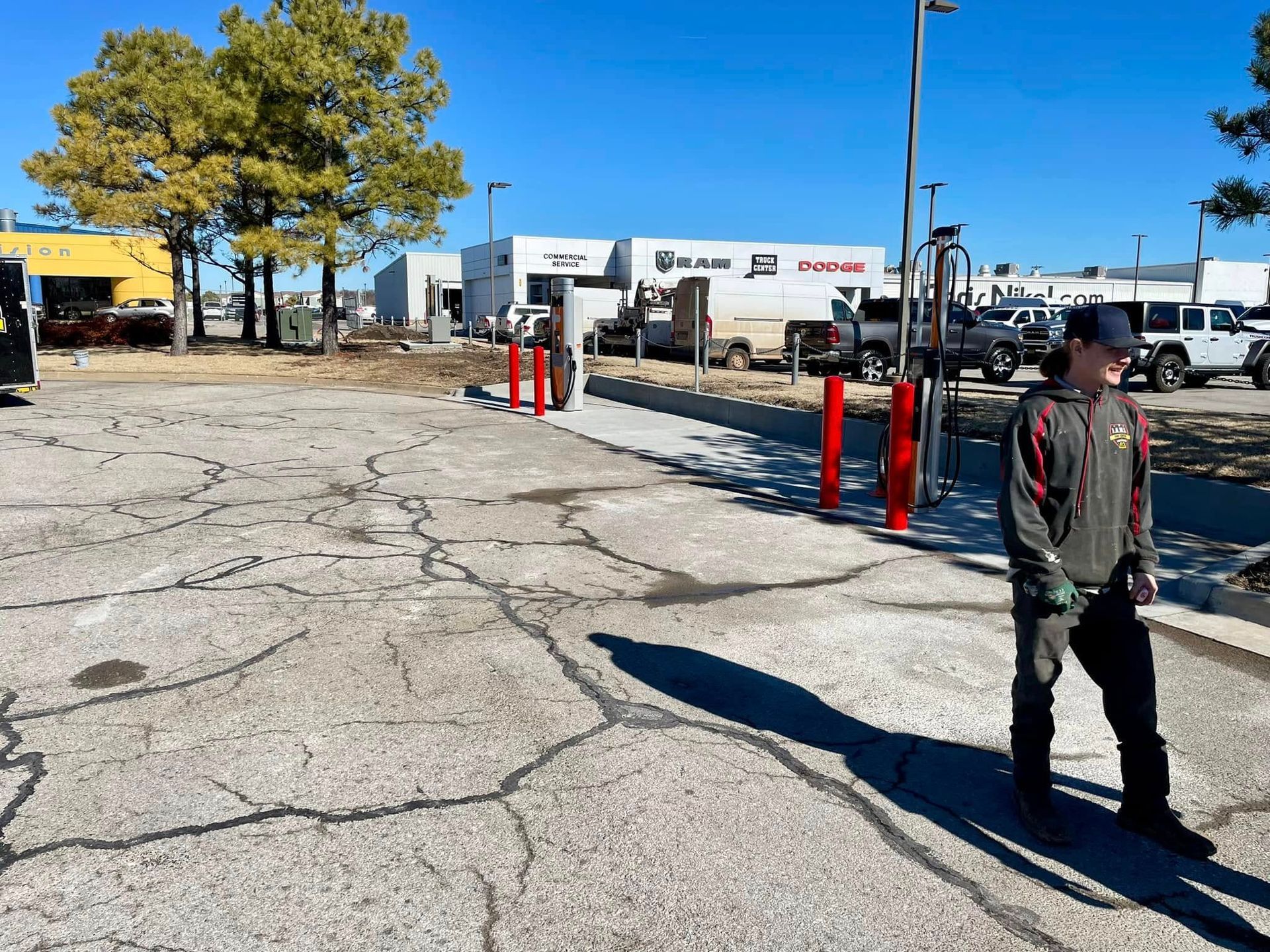 Person walking in a cracked parking lot near red bollards and trees under a clear blue sky