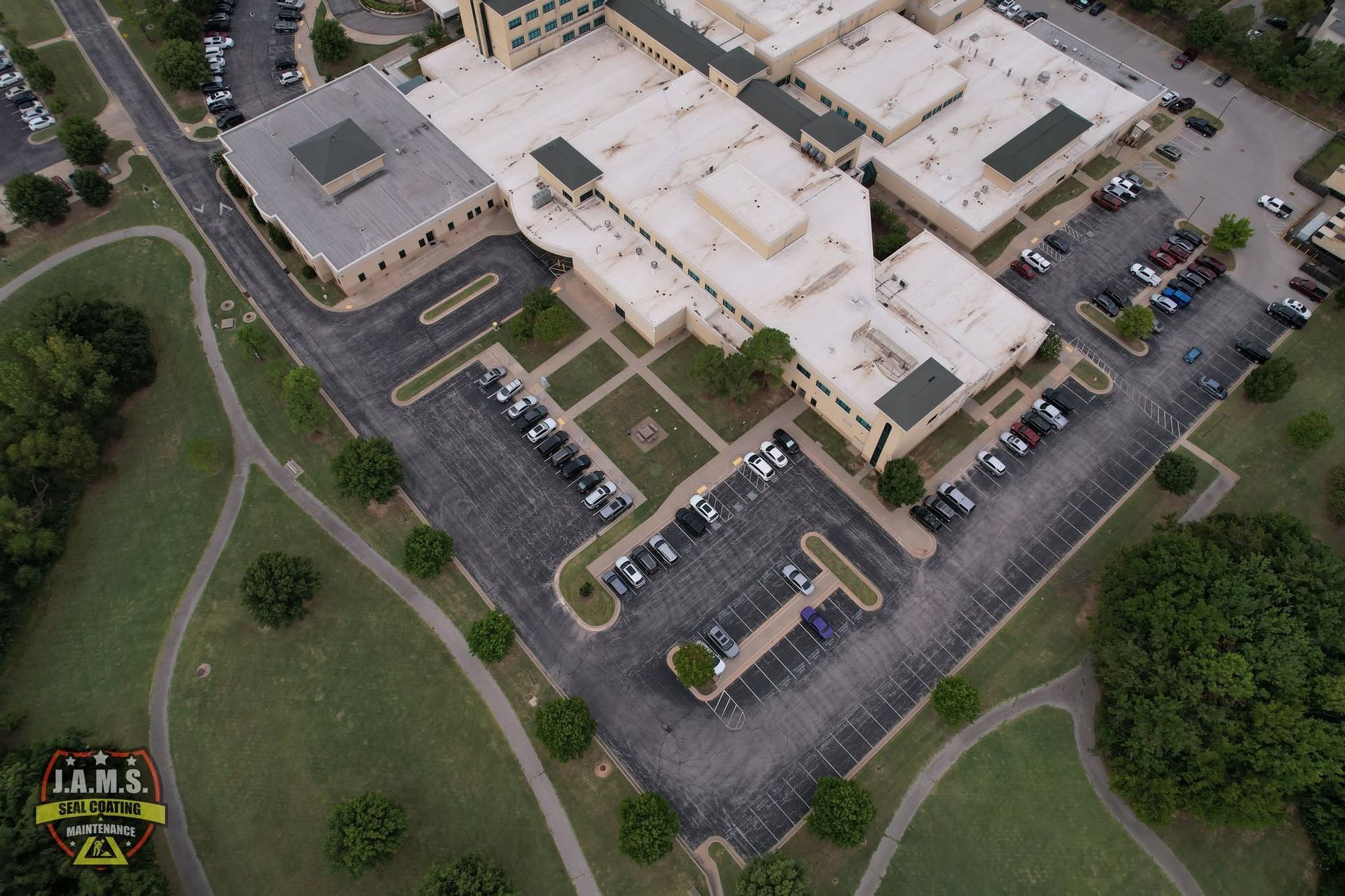 Aerial view of a large beige building with parking lots, roads, and surrounding green trees.