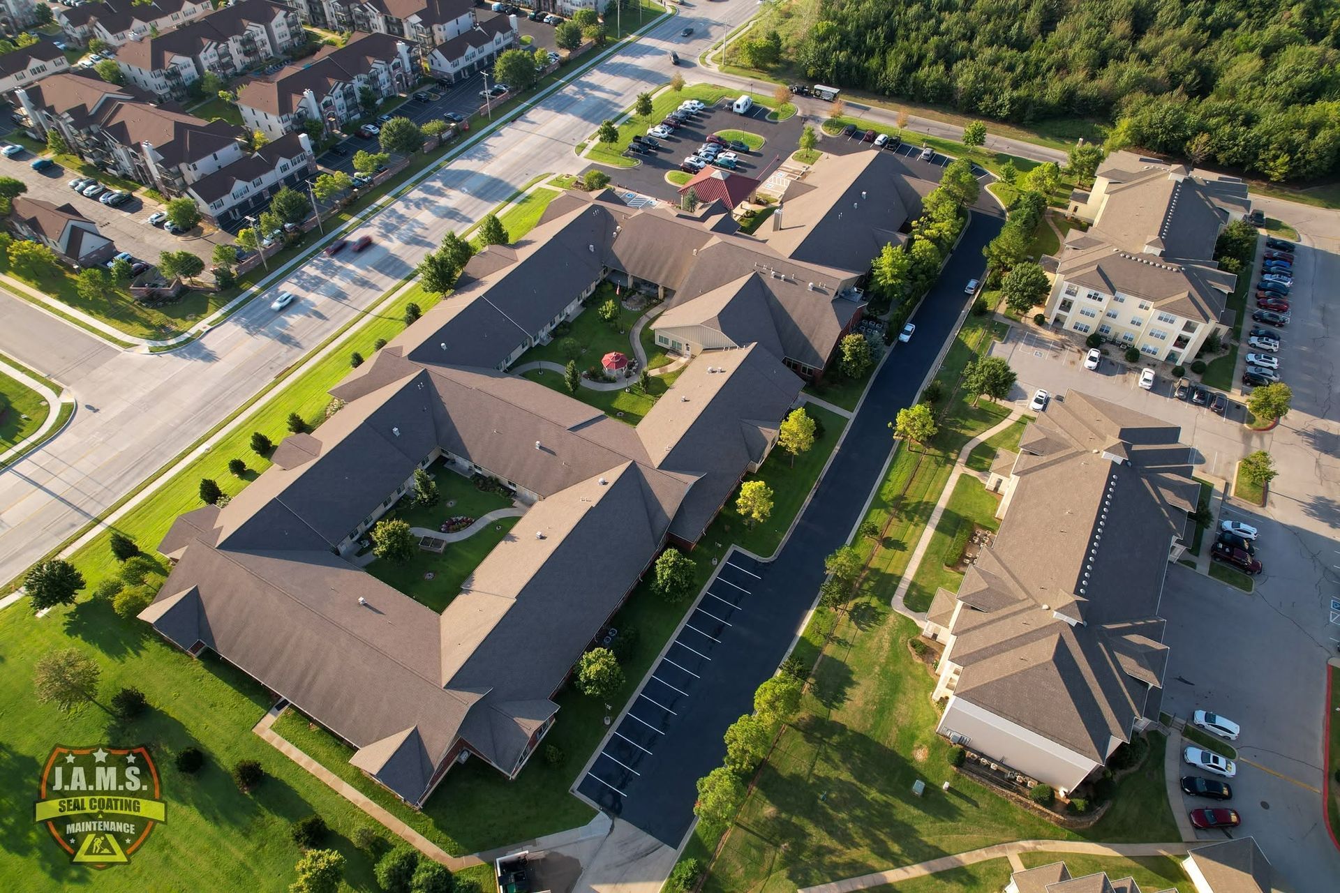 Aerial view of a beige apartment complex with parking lots, roads, and surrounding homes on a sunny day