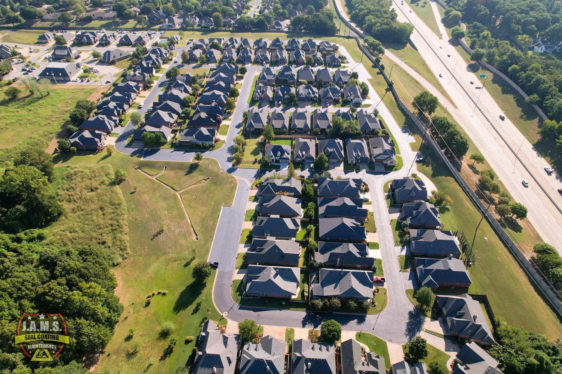 Aerial view of a suburban neighborhood with rows of dark-roofed houses beside a highway and green open space