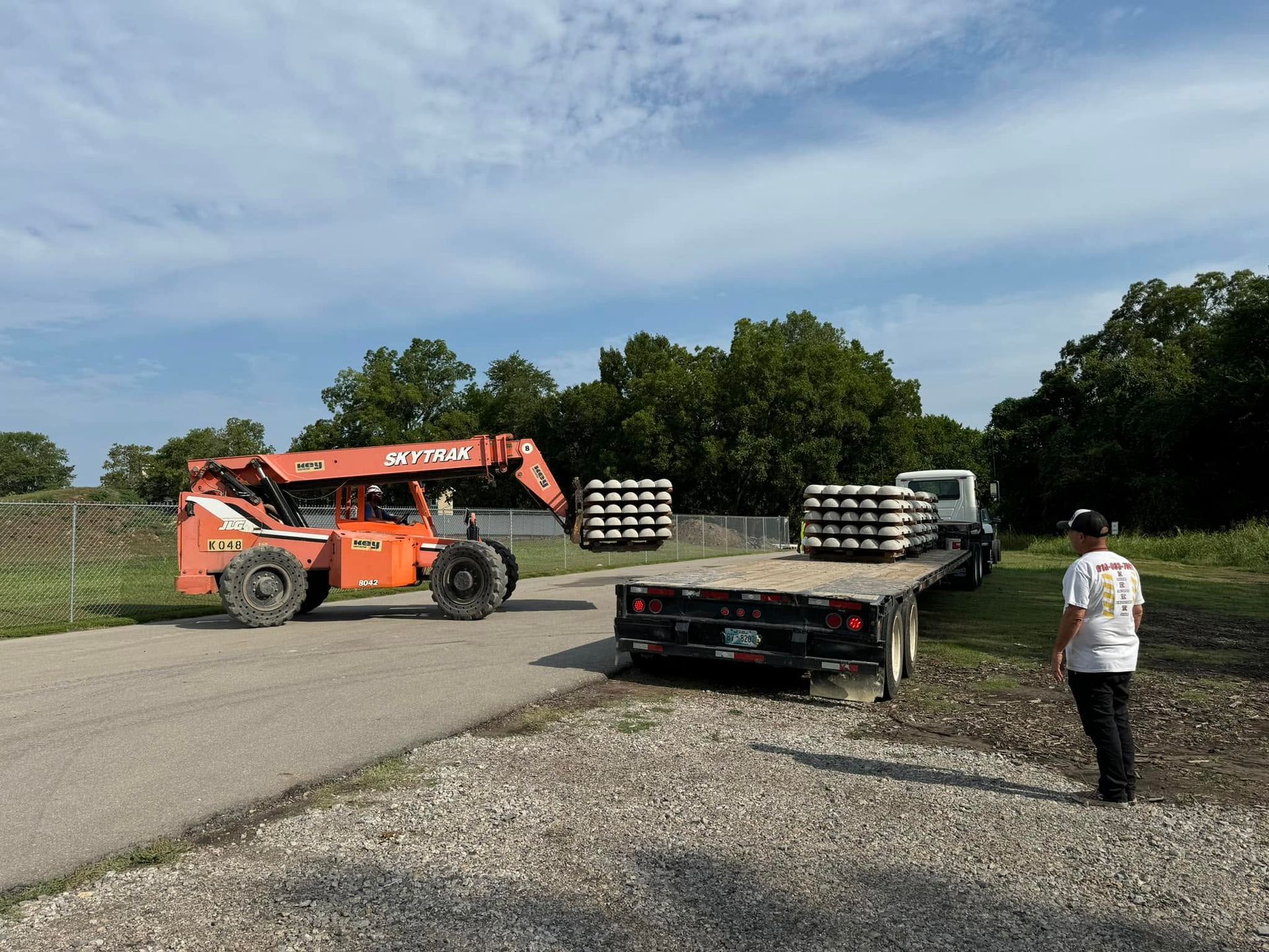 Orange forklift loading stacked pallets onto a truck in a gravel lot, with a person standing nearby.