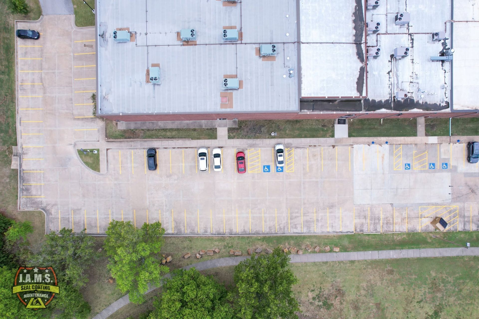 Aerial view of a mostly empty parking lot beside a building with a few parked cars and trees nearby.