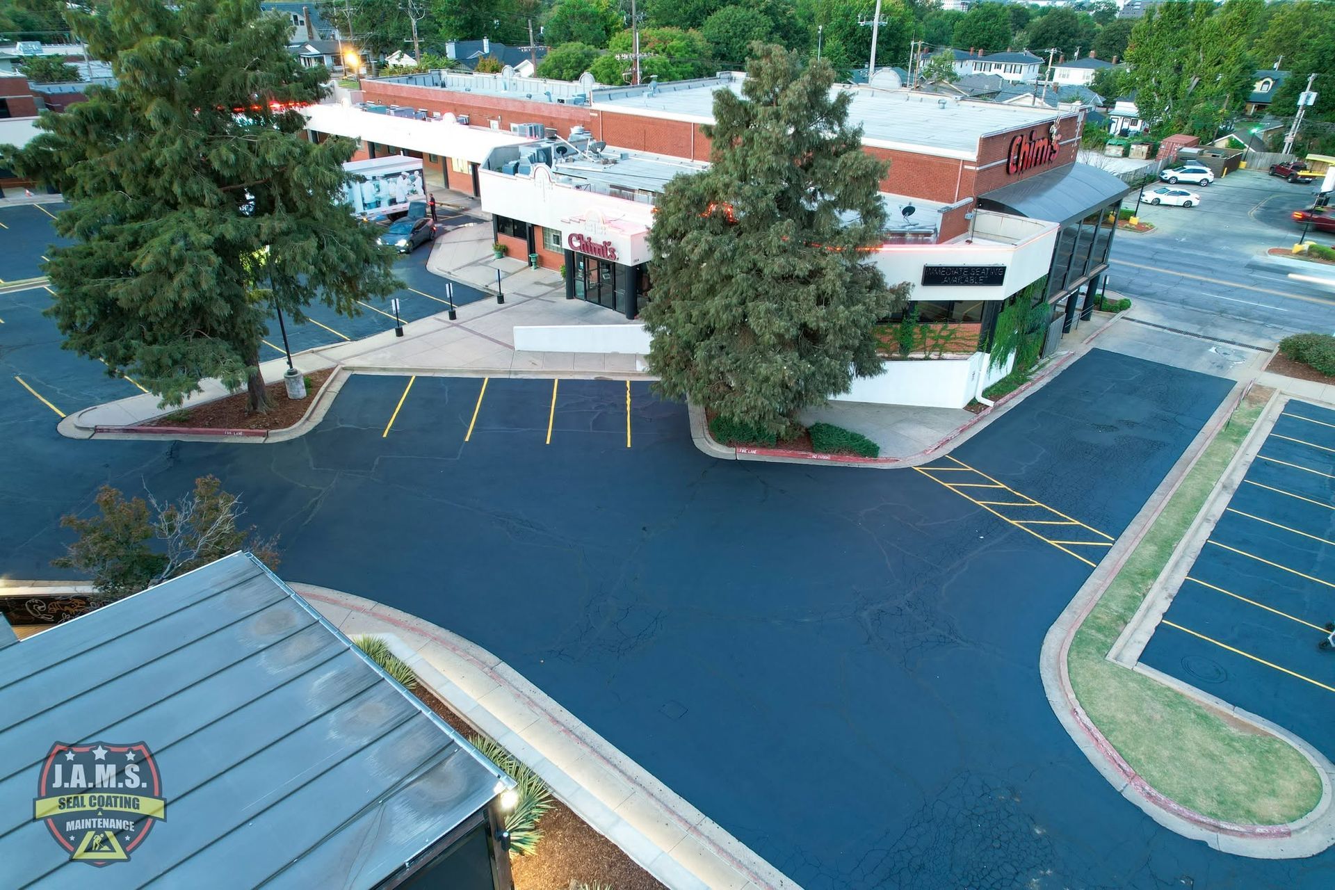 Aerial view of a strip mall parking lot with storefronts, trees, and mostly empty spaces.