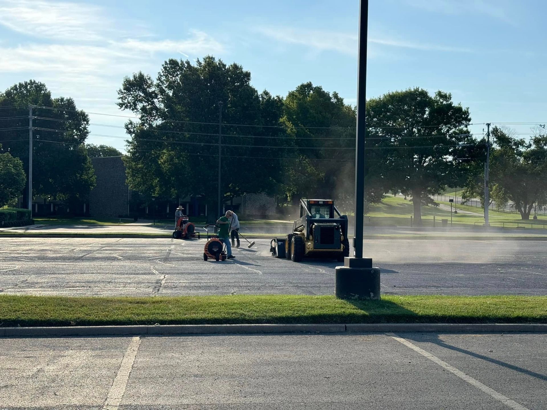 Small tractor spraying dust on an empty parking lot near a few people and trees in the distance