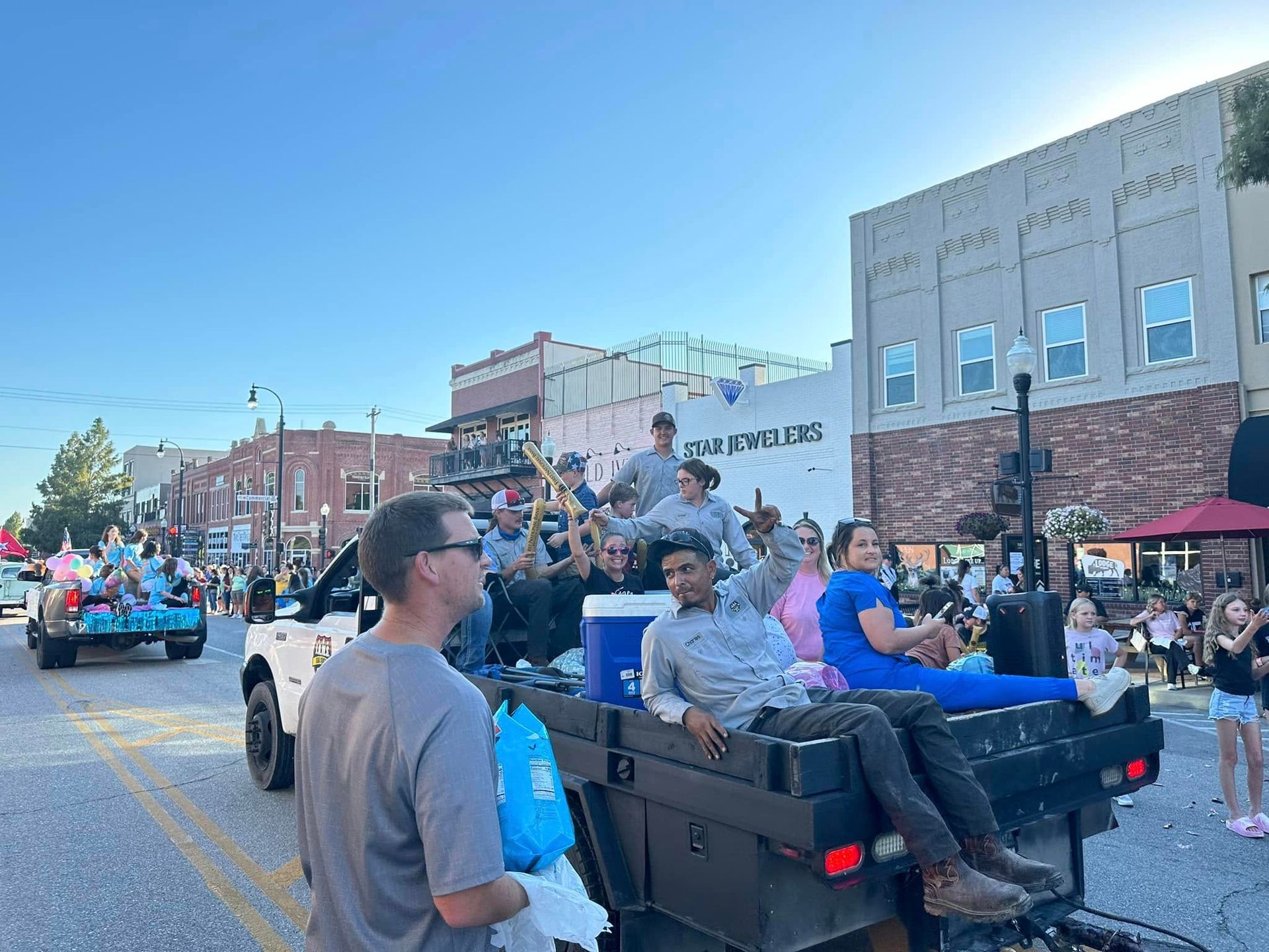 Parade float with people riding in the back of a black pickup, crowd lining the street under a clear sky.