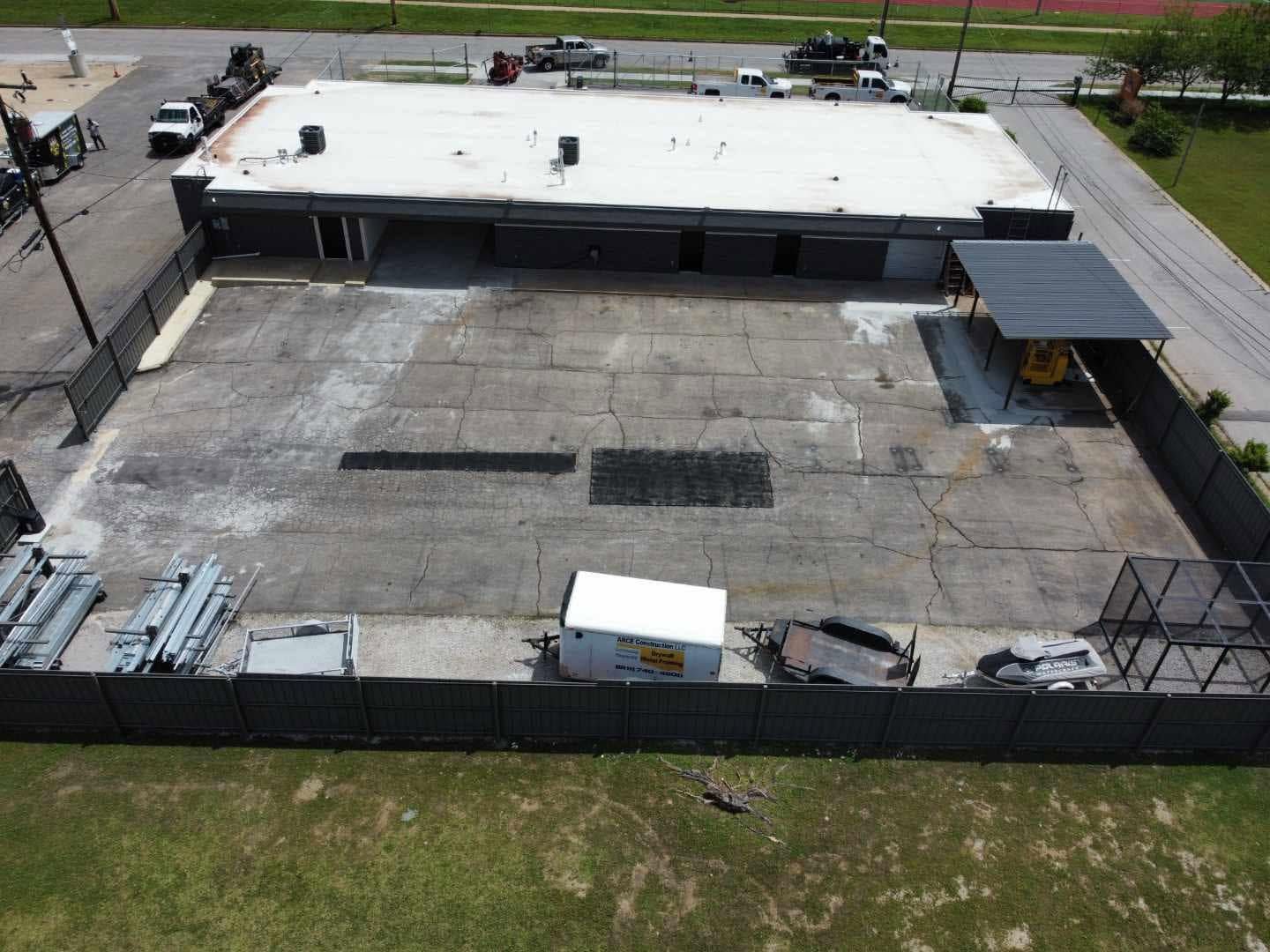 Aerial view of a fenced industrial lot with a flat-roofed building and scattered equipment.