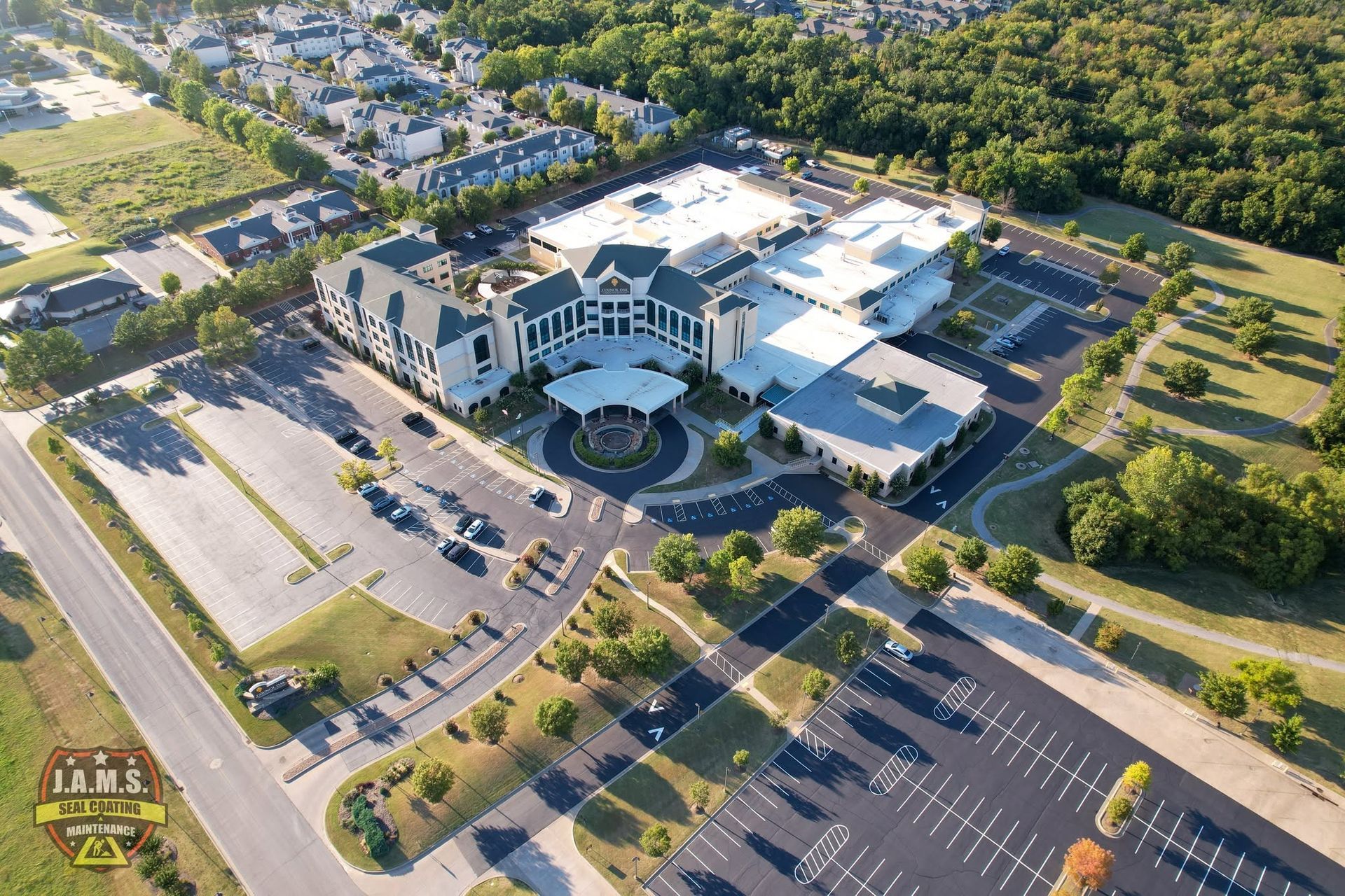 Aerial view of a large building complex with parking lots, roads, and surrounding trees in daylight