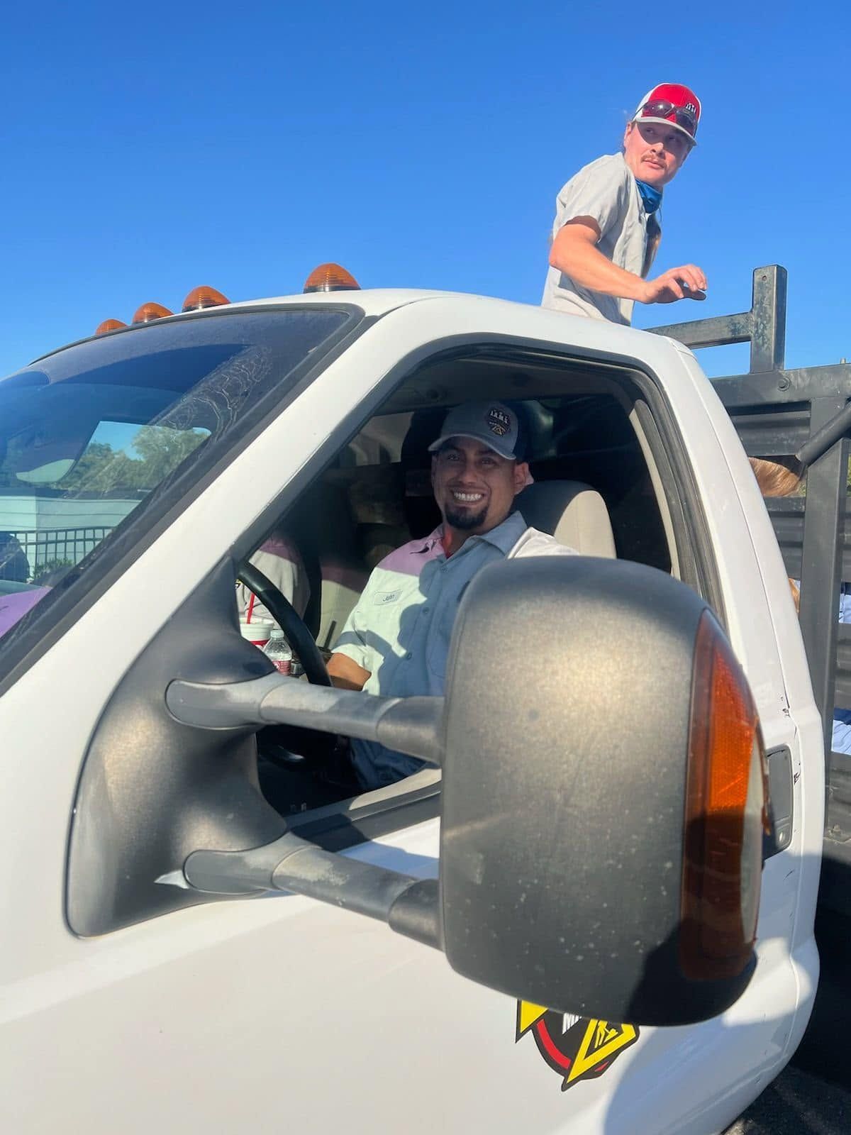 Person in a white pickup truck smiles while another stands on the roof in a red cap under a blue sky