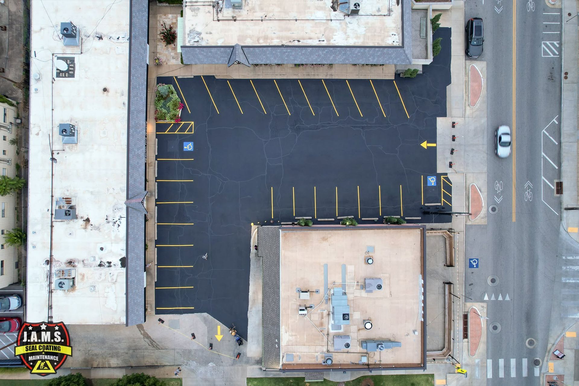 Aerial view of an empty parking lot between buildings and a street with parked cars.
