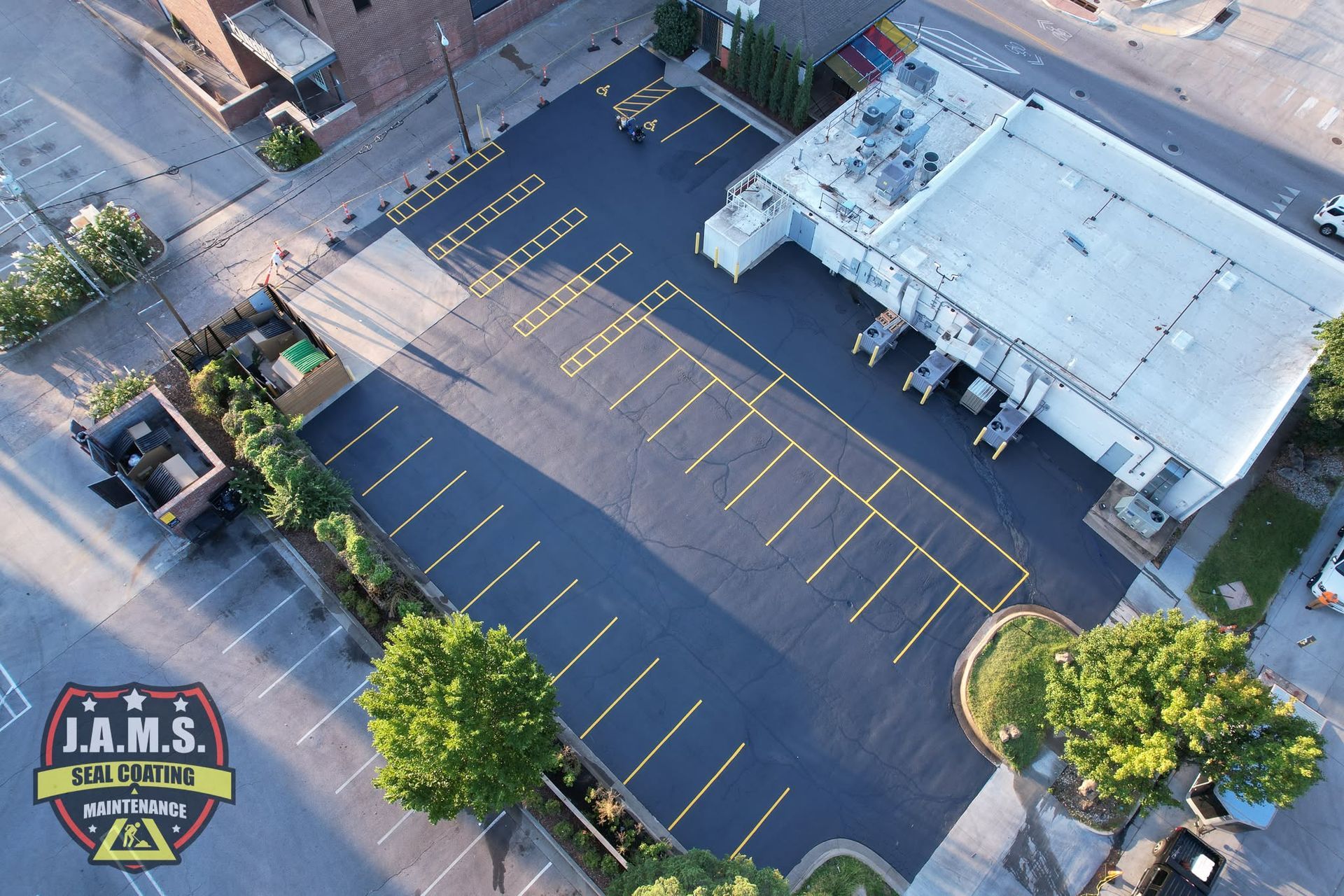 Aerial view of a mostly empty parking lot beside a white-roofed building and a J.A.M.S. sign at bottom left