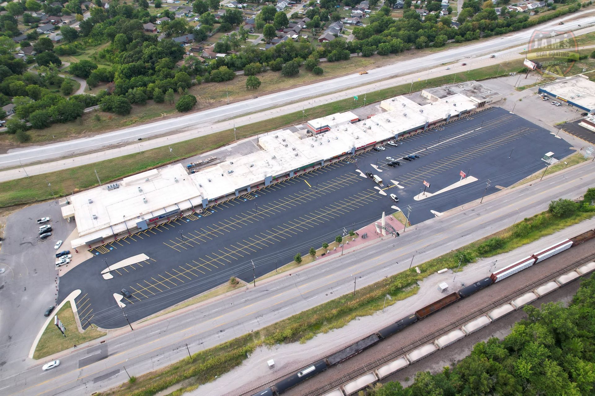 Aerial view of a large parking lot beside a long white-roofed building and a road near trees.